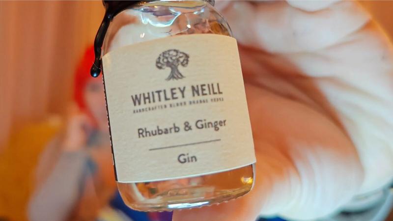 Photo: A hand holding a small glass bottle of gin towards the camera in sharp focus against a warm, softly blurred background. The bottle has a black screw cap and a cream-coloured label featuring a stylised tree illustration at the top, followed by "WHITLEY NEILL" in bold text with smaller text beneath reading "ESTABLISHED IN 1762 WIRRAL ESSEX", then "Rhubarb & Ginger" as the main descriptor, a horizontal line, and "Gin" at the bottom. The bottle contains pale golden or pink-tinted liquid. The background shows warm peachy-orange tones with soft focus, suggesting an interior setting with warm lighting. A hint of red is visible on the left edge, and indistinct blurred shapes suggest people in the background.