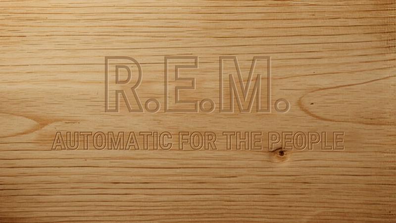 A close-up of warm pale wood grain, with "R.E.M." and "AUTOMATIC FOR THE PEOPLE" pressed into the surface as if carved there. The kind of thing you might find on a shelf, slightly forgotten, until you need it.