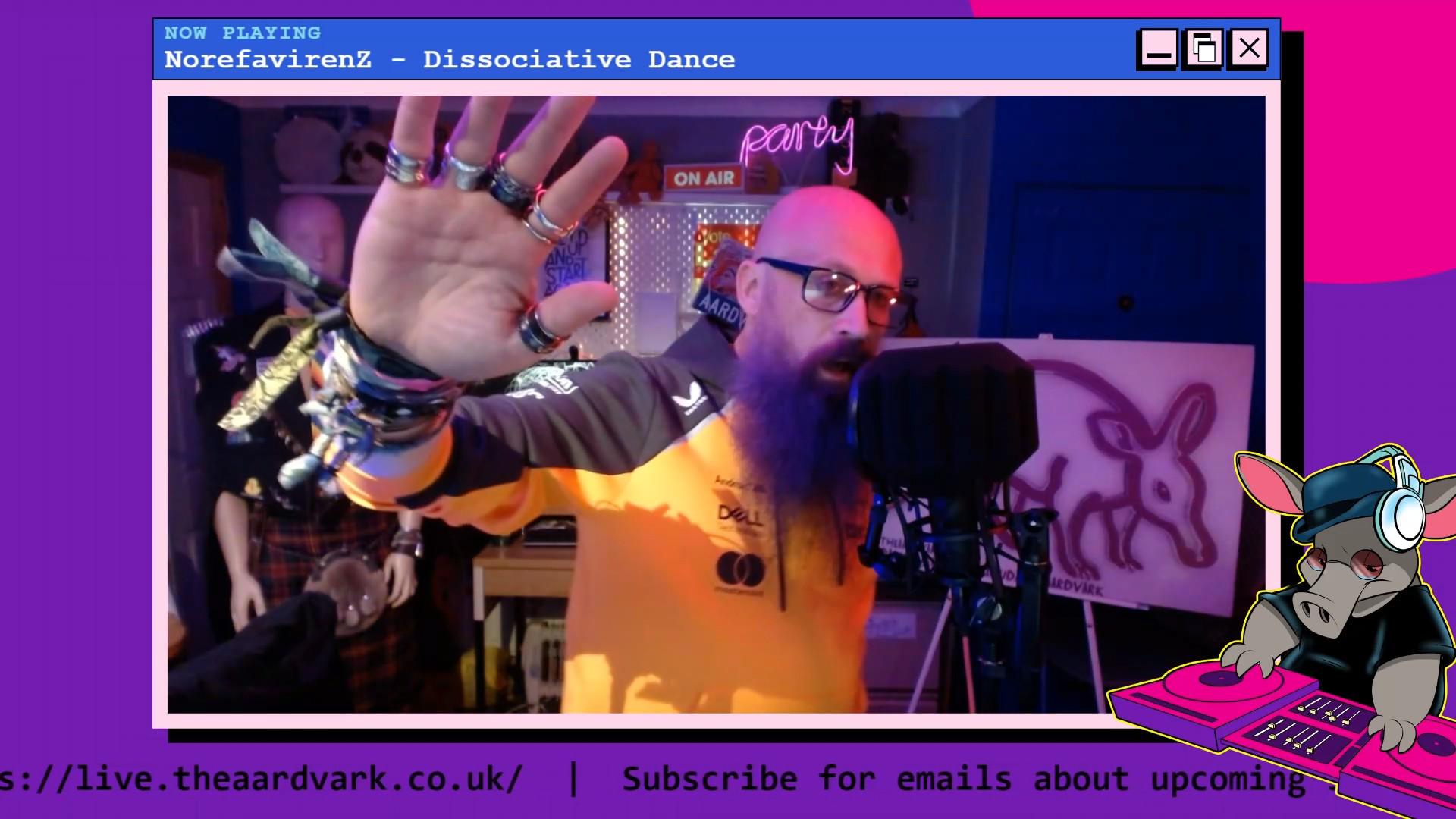 Screenshot from theaardvark live, captured by Winni from Germany. A bald, bearded man in an orange top raises a heavily-ringed hand toward the camera with the energy of someone making an extremely important point about a track called "Dissociative Dance." The NOW PLAYING panel confirms this is exactly what is happening. Behind him, a mannequin in a suit and kilt stands in its usual position of silent judgement. The aardvark show banner is partially visible on the right. The "ON AIR" sign is lit. The aardvark DJ character in the bottom right corner appears to be the most focused person in the room.