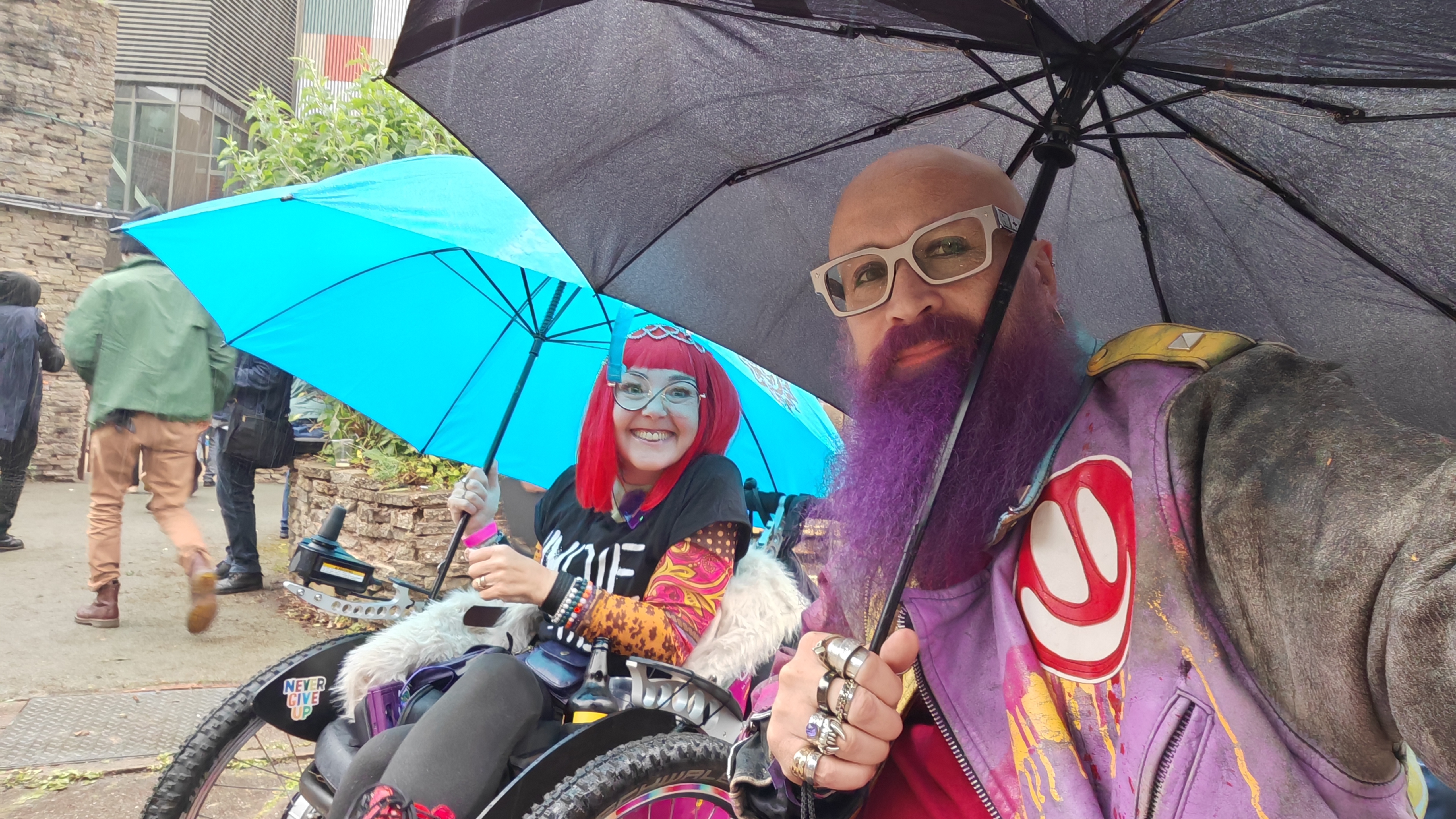 Two people shelter under separate umbrellas in the rain outside the Midlands Arts Centre. On the left, Sha holds a cyan umbrella and grins at the camera with the expression of someone who has decided the weather is not going to be the boss of her. On the right, a bald man with a large purple beard and white-framed glasses holds a black umbrella and a look of mild resignation, wearing a purple jacket with a large red smiley face that appears to be melting. A "Never Give Up" sticker is visible on Sha's wheelchair. They clearly have the energy of people who've only just arrived.