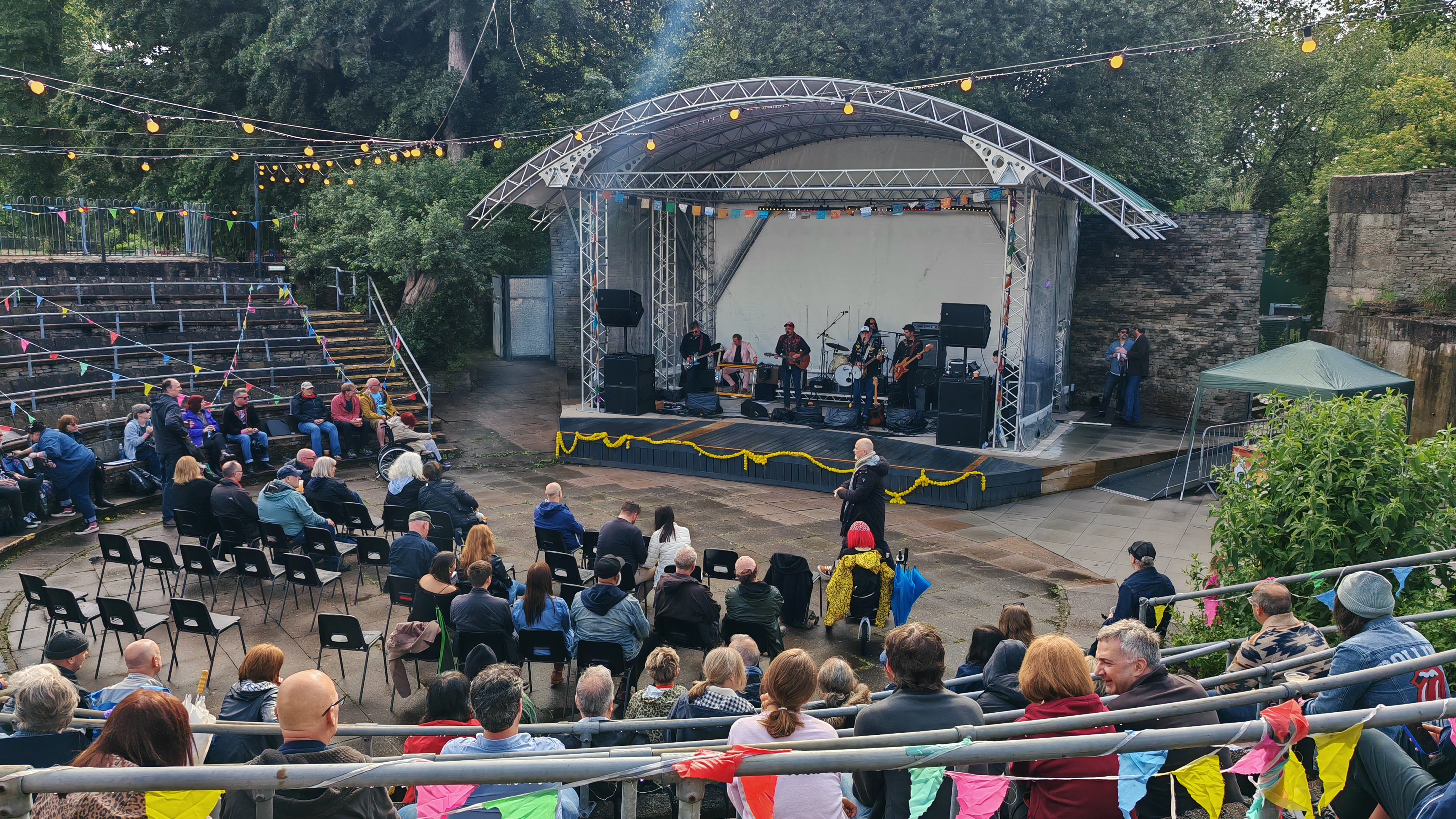 The outdoor amphitheatre at the Midlands Arts Centre, shot from the top of the stone terraced seating. Independent Country are on stage beneath a curved steel roof, bunting strung across the venue, fairy lights waiting for the evening. The audience fills the terraces and the standing area below — coats on, a few umbrellas still up. Near the front of the standing area, a figure in a bright yellow poncho with red hair is visible, which is either Sha or a very committed lighthouse. The trees behind the stage are indifferent to all of this.
