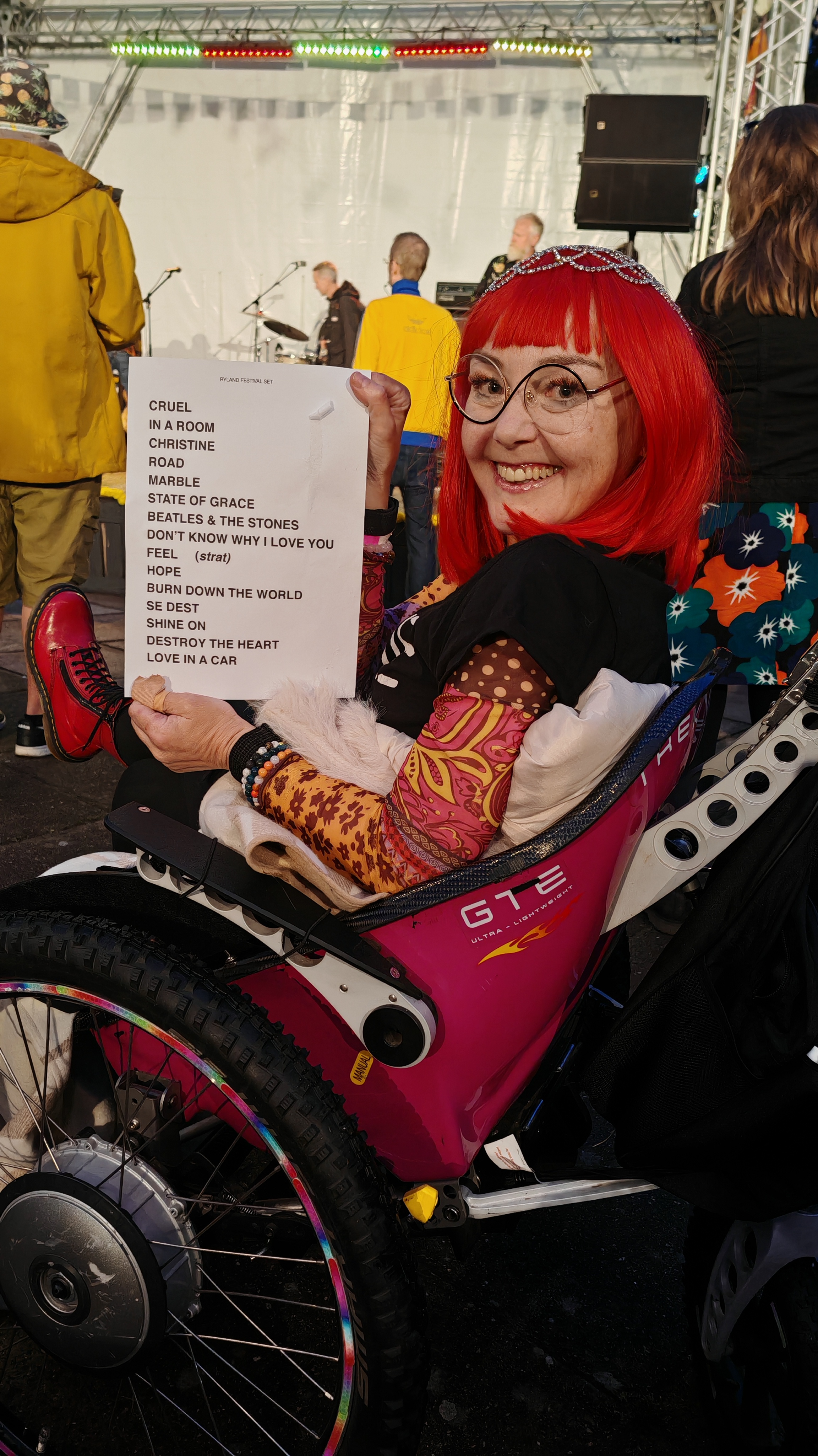 A woman with red hair, jewelled hairnet and round glasses sits in her pink wheelchair in front of an outdoor stage, holding up a printed setlist for the Ryland Festival set with the expression of someone who has just won something. The setlist reads: Cruel, In a Room, Christine, Road, Marble, State of Grace, Beatles & The Stones, Don't Know Why I Love You, Feel (strat), Hope, Burn Down The World, Se Dest, Shine On, Destroy The Heart, Love In A Car. A tech from the MAC grabbed it for her from the stage. Behind her, the band are still on stage and the audience are still watching. The setlist had been identified as a target approximately four hours earlier. No one else stood a chance.