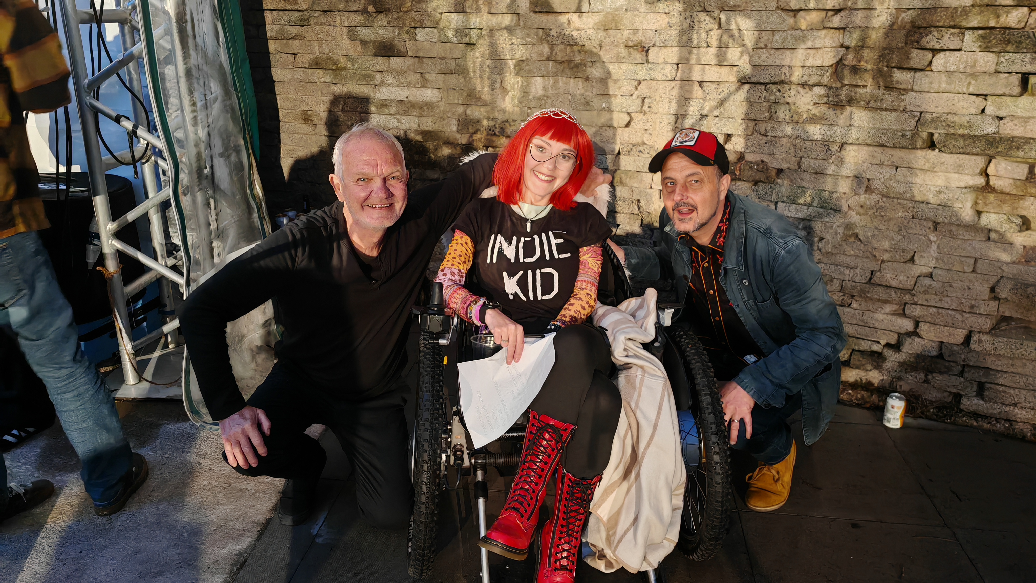 Three people pose against a stone wall backstage at a music festival. On the left, Guy Chadwick of The House of Love crouches in all black, smiling with the easy warmth of someone who has been doing this for forty years. On the right, Craig from Independent Country crouches in a denim jacket, doing the same. In the middle, Sha sits in her wheelchair wearing an "Indie Kid" t-shirt, a jewelled hairnet, and red DM boots, holding a sheet of interview questions she absolutely intended to use. All three are grinning. The two men are crouching to be level with her. She is the tallest and most ecstatic person in the photo.