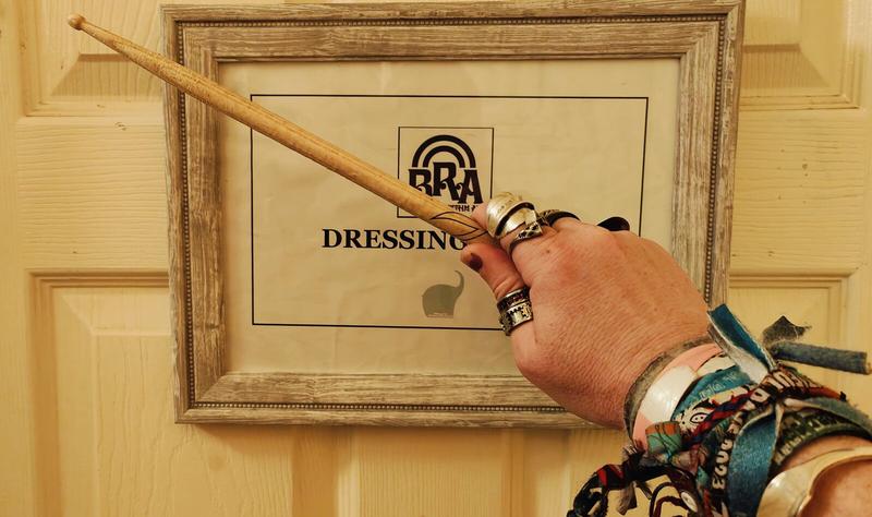 My right hand, wearing too many rings and festival wristbands, holding a drumstick. Behind it is a framed sign on a door. The sign has Bentley Rhythm Ace's logo and the partially obscured words "Dressing Room"