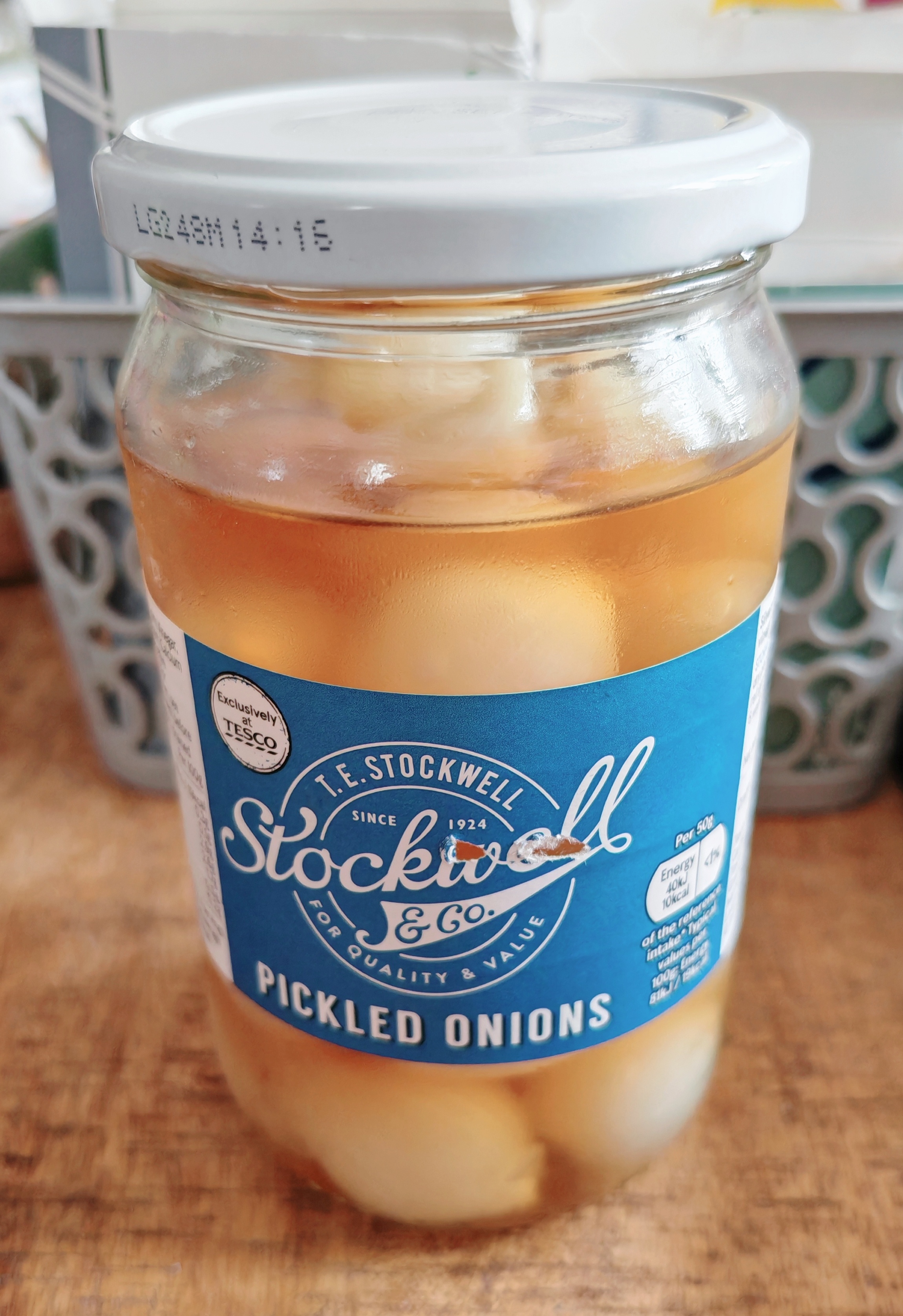 A glass jar of Stockwell & Co pickled onions sits on a wooden worktop, looking about as enthusiastic as the onions inside - which appear rather anaemic and floating listlessly in their "vinegar and acetic acid" like they've given up on life. The blue label promises quality since 1924, though the pale, unappetising contents suggest standards may have slipped a touch.