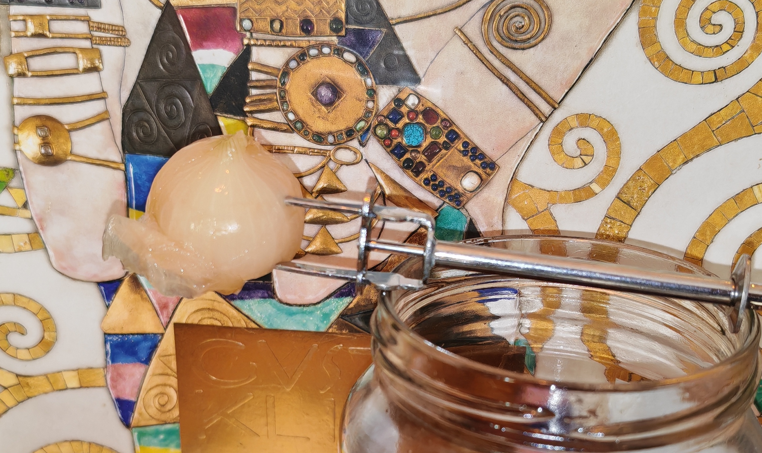 A pale, translucent pickled onion sits impaled on a small silver cocktail fork, balanced precariously across the rim of its glass jar like a reluctant diver contemplating the plunge back into the amber depths below. The fork's metallic surface catches the light against the sumptuous backdrop of Klimt's swirling golden spirals and jewel-encrusted mosaics - rather like presenting a modest pub snack on a Renaissance altar. The onion itself appears to have accepted its fate with typical British stoicism.