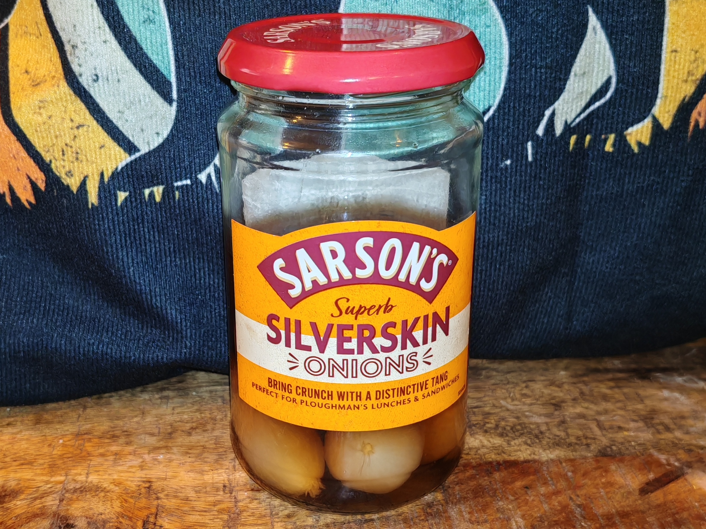 A jar of Sarson's Superb Silverskin onions sits on a wooden surface against a colourful fabric backdrop featuring bold red, orange, yellow, and turquoise stripes. The orange and burgundy label promises "bring crunch with a distinctive tang" - ambitious claims for what is a merely acceptable addition to a pickled onion enthusiast's pantry.