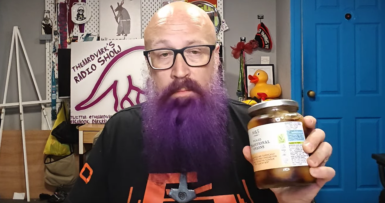 A bald, bespectacled chap with an impressively purple-tinted beard holds up a jar of M&S pickled traditional onions to the camera. Behind him sits the controlled chaos of a home office-cum-recording studio, complete with a "theaardvark's Radio Show" poster, a rubber duck (because of course there is), and a bright blue door that's probably seen more drama than EastEnders. The expression suggests this pickle jar might contain either culinary gold or his greatest mistake - the sort of face one pulls when about to find out if yesterday's leftover curry was a brilliant idea or a terrible miscalculation.