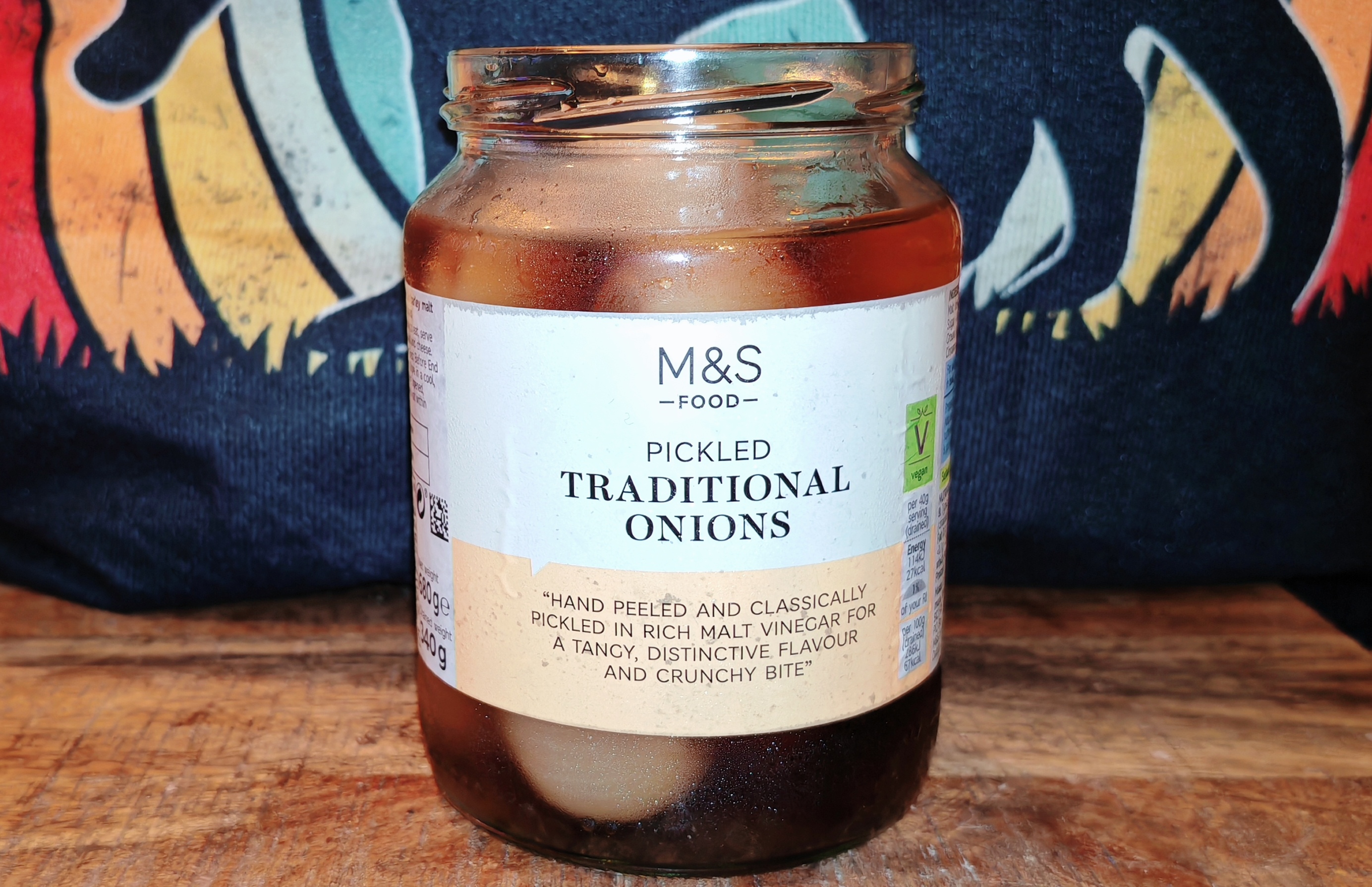 A glass jar of M&S pickled traditional onions sits on a wooden surface against a backdrop of vibrant knitted fabric in stripes of red, orange, yellow, navy, and mint green. The jar contains golden-brown vinegar with pale pickled onions visible through the glass, while the white and cream label prominently displays the M&S Food branding and promises these are "hand peeled and classically pickled in rich malt vinegar for a tangy, distinctive flavour and crunchy bite.