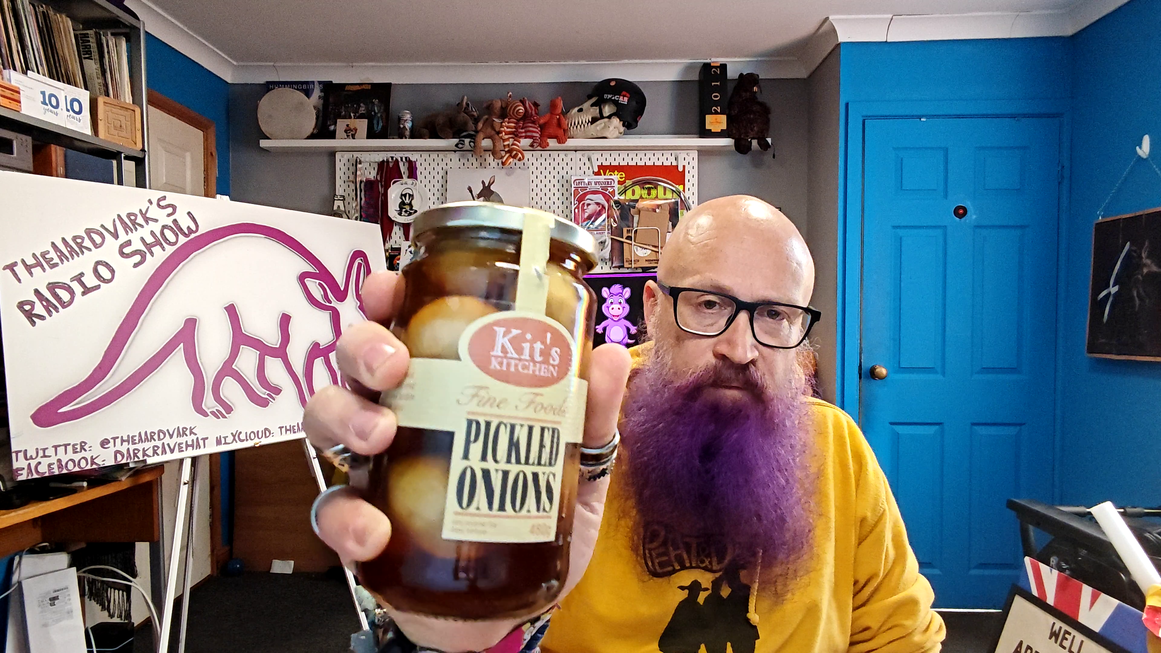 Bald man with glasses and a magnificent purple-dyed beard wearing a mustard yellow hoodie, holding up a jar of Kit's Kitchen pickled onions towards the camera in his home studio. Behind him are shelves crammed with collectibles, a bright blue door, and a white banner reading "THEAARDVARKS RADIO SHOW" with a pink cartoon aardvark logo. The pickled onions look suspiciously appetising for something that's about to be thoroughly judged.