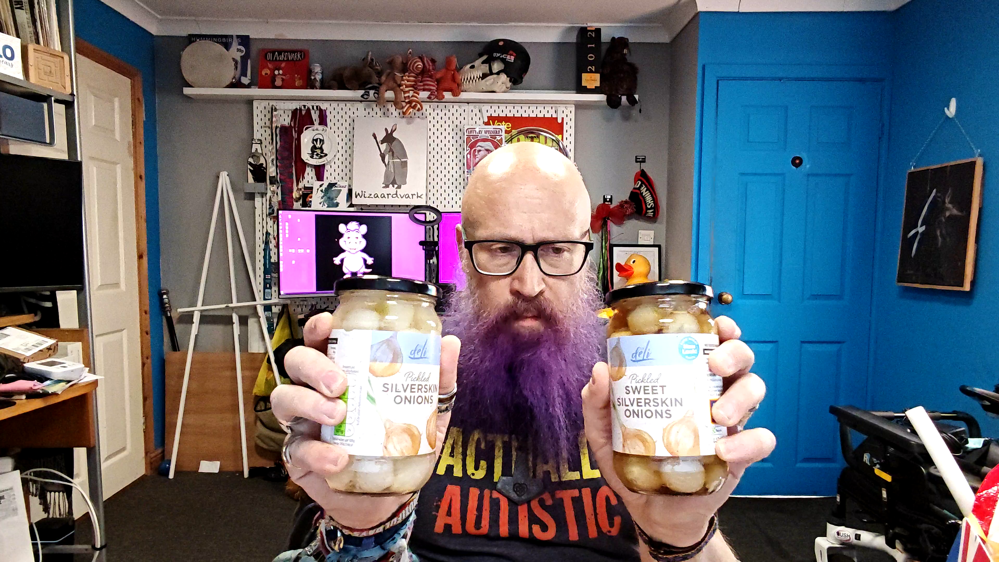 Bald man with glasses and a purple beard wearing a grey t-shirt with "ACTUALLY AUTISTIC" in yellow and orange text, holding up two jars of pickled silverskin onions - one labelled "Pickled Silverskin Onions" and the other "Pickled Sweet Silverskin Onions". Behind him is a cluttered home office with shelves of collectibles, a bright blue door, and computer monitors displaying a purple cartoon aardvark. The expression suggests he's about to deliver a verdict that could make or break someone's onion-pickling career.