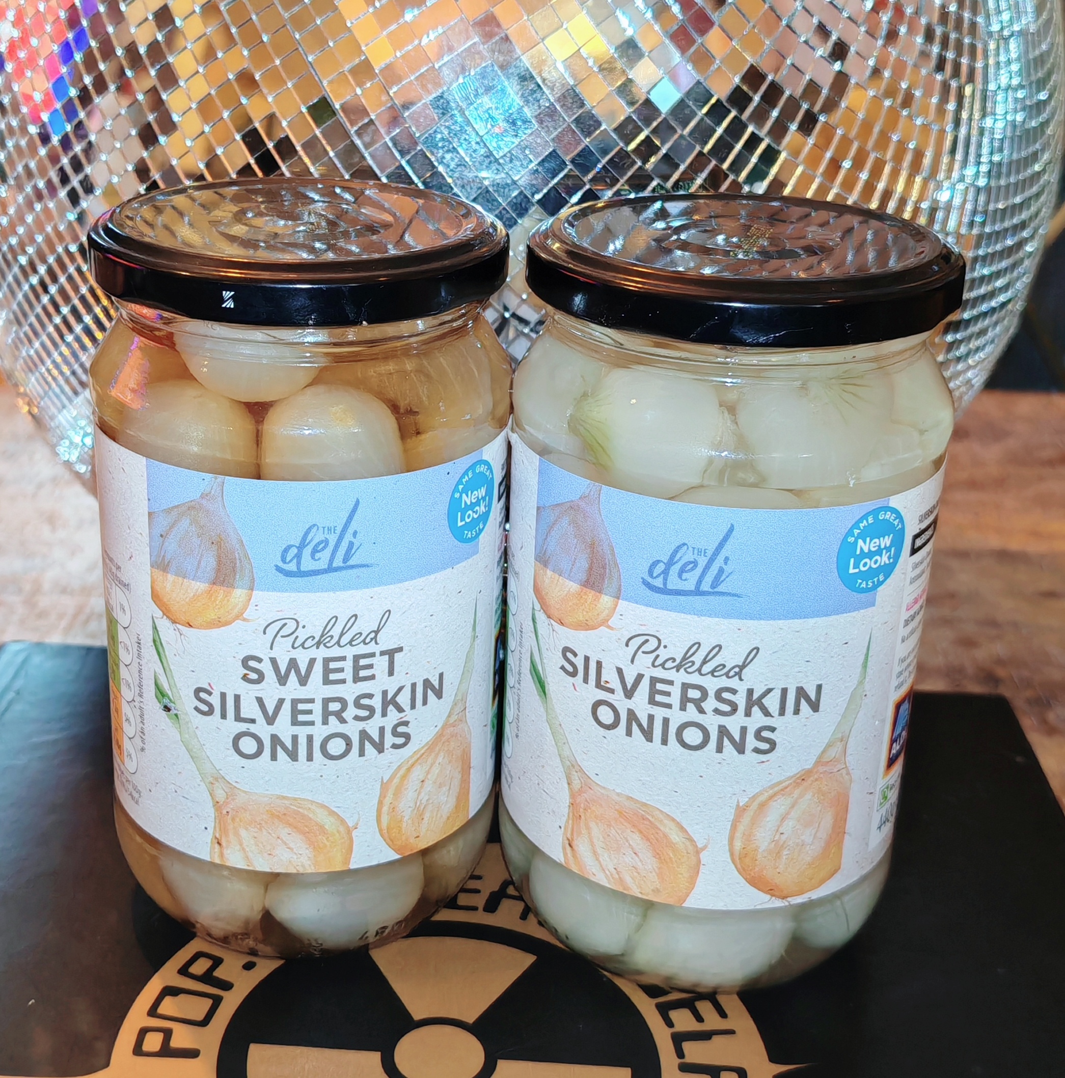 Two glass jars of pickled silverskin onions from Aldi's Deli range sit on a dark wooden surface against a shimmering silver disco ball backdrop. The left jar contains "Sweet Silverskin Onions" with tiny onions in a dirty brown liquid, whilst the right jar holds plain "Silverskin Onions" with pale white onions. Both have black lids and white labels featuring illustrated onions.