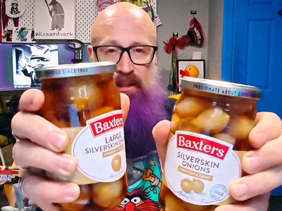 Bald man with glasses and a purple beard holding up two jars of Baxters pickled silverskin onions - on the left "Large Silverskin Onions" and on the right "Silverskin Onions"  - both with distinctive red and white Baxters labels. The jars are held close to the camera, showing the onions floating in their brown pickling liquid. Behind him is his disorganised home office setup with collectibles on shelves, including what appears to be a rubber duck and various pop culture memorabilia. His expression suggests he's mid-explanation, presumably about the merits of Scotland's finest preserved alliums.