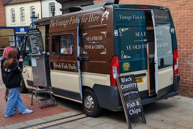 Photo: Mobile fish and chip van called "The Vintage Fish and Chips Van" parked on a high street, with brown and teal livery advertising "Sea Fresh Fish & Chips" and "Fresh Fish Daily". A customer in jeans stands at the serving window, and there's a pavement sign advertising "Black Country" fish & chips for £6.50.