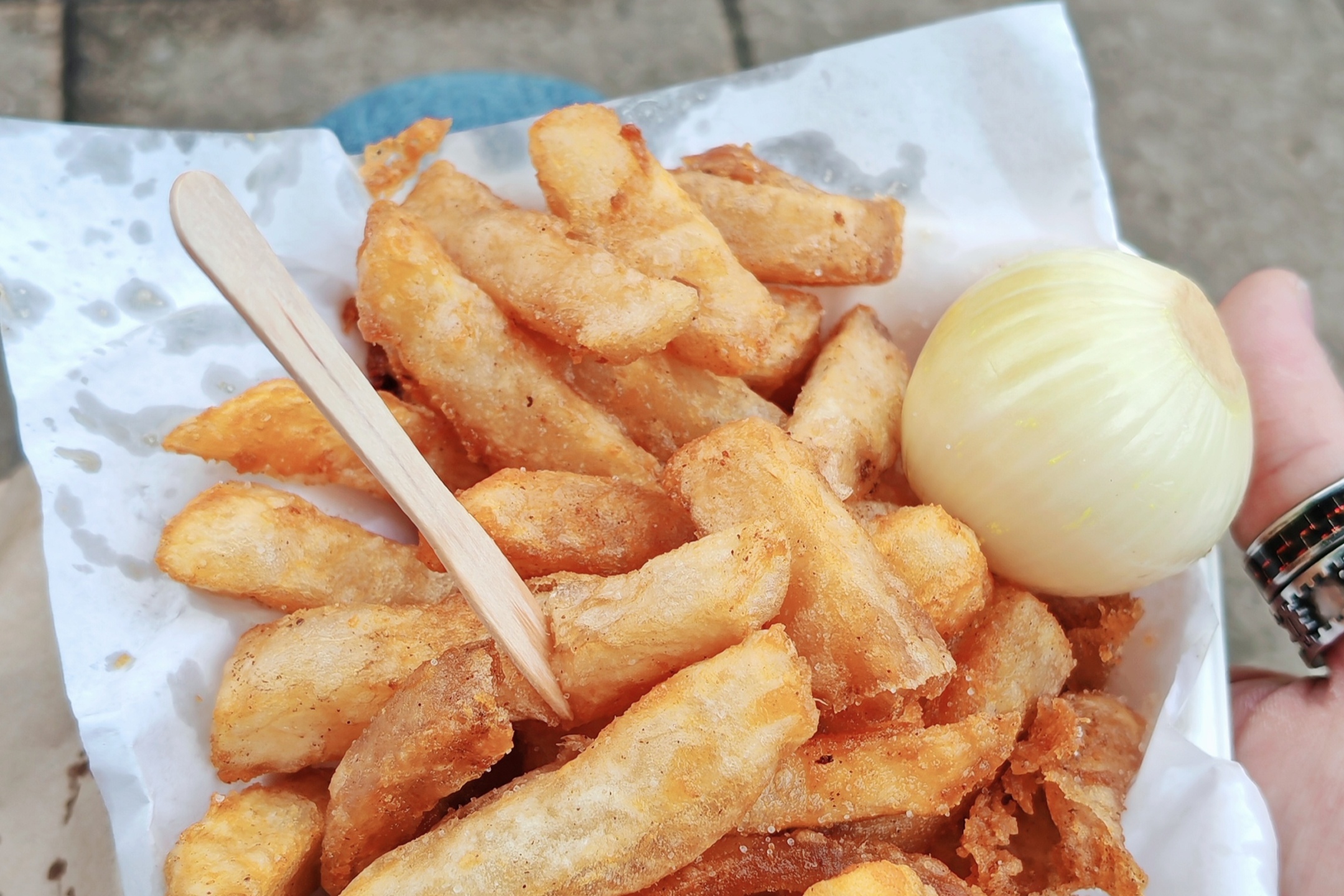 Photo: Hand holding a portion of golden chips in white paper wrapping with a wooden fork, accompanied by a pickled onion. The chips look properly crispy and well-seasoned, photographed with a stone surface background. Classic Black Country fare at its finest.