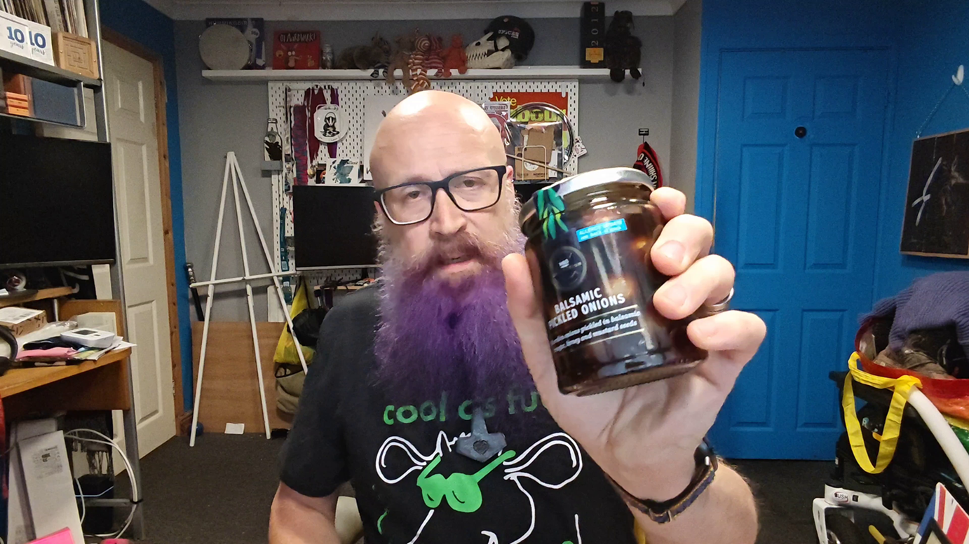 Screen grab: Bald man with glasses and a purple beard wearing a black Inspiral Carpets t-shirt with green text reading "cool as fuck" and a cow wearing green sunglasses, holding up a jar of balsamic pickled onions towards the camera. Behind him is his familiar home office with shelves of collectibles, a bright blue door, and various pop culture memorabilia. His expression suggests he's about to deliver another informed verdict on the quality of preserved alliums.