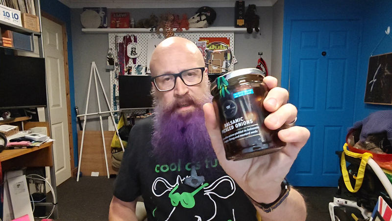 Screen grab: Bald man with glasses and a purple beard wearing a black Inspiral Carpets t-shirt with green text reading "cool as fuck" and a cow wearing green sunglasses, holding up a jar of balsamic pickled onions towards the camera. Behind him is his familiar home office with shelves of collectibles, a bright blue door, and various pop culture memorabilia. His expression suggests he's about to deliver another informed verdict on the quality of preserved alliums.