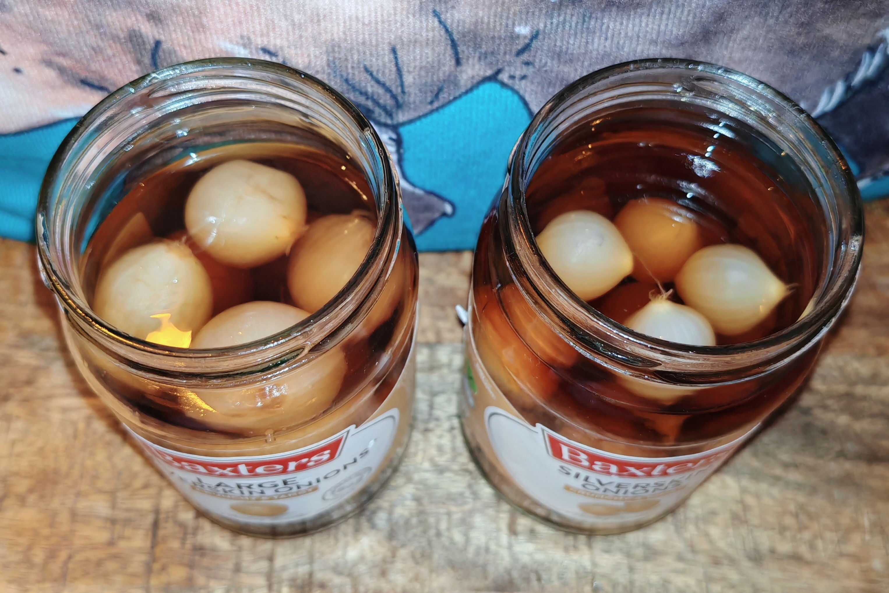 The same two Baxters jars now opened, showing a top-down view of the pickled onions floating in their dark amber pickling liquid. The onions appear plump and well-preserved, with the liquid having that rich, malty colour that suggests proper fermentation. The aardvark print provides a rustic backdrop as the evaluation begins.