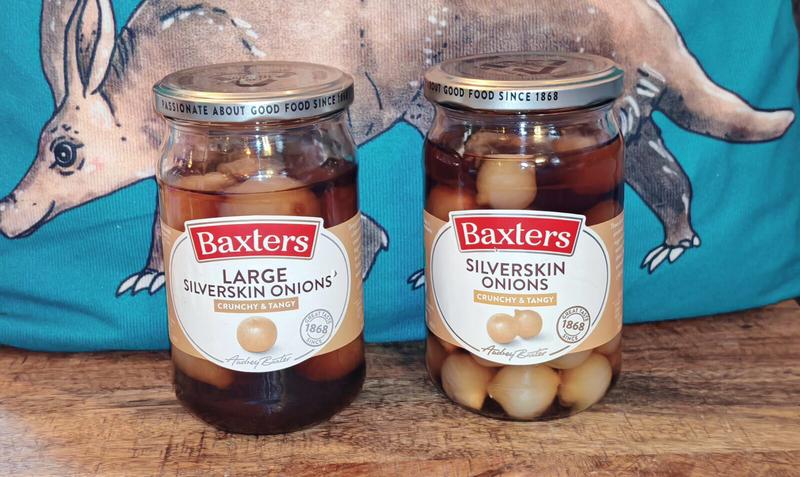 Photo: Two sealed jars of Baxters pickled onions sitting side by side on a wooden surface - one large silverskin onions, one regular silverskin onions - with an aardvark print on turquoise material as backdrop. Both jars display the classic Baxters branding with "Passionate About Good Food Since 1868" on the lids, ready for the tasting that's about to commence.