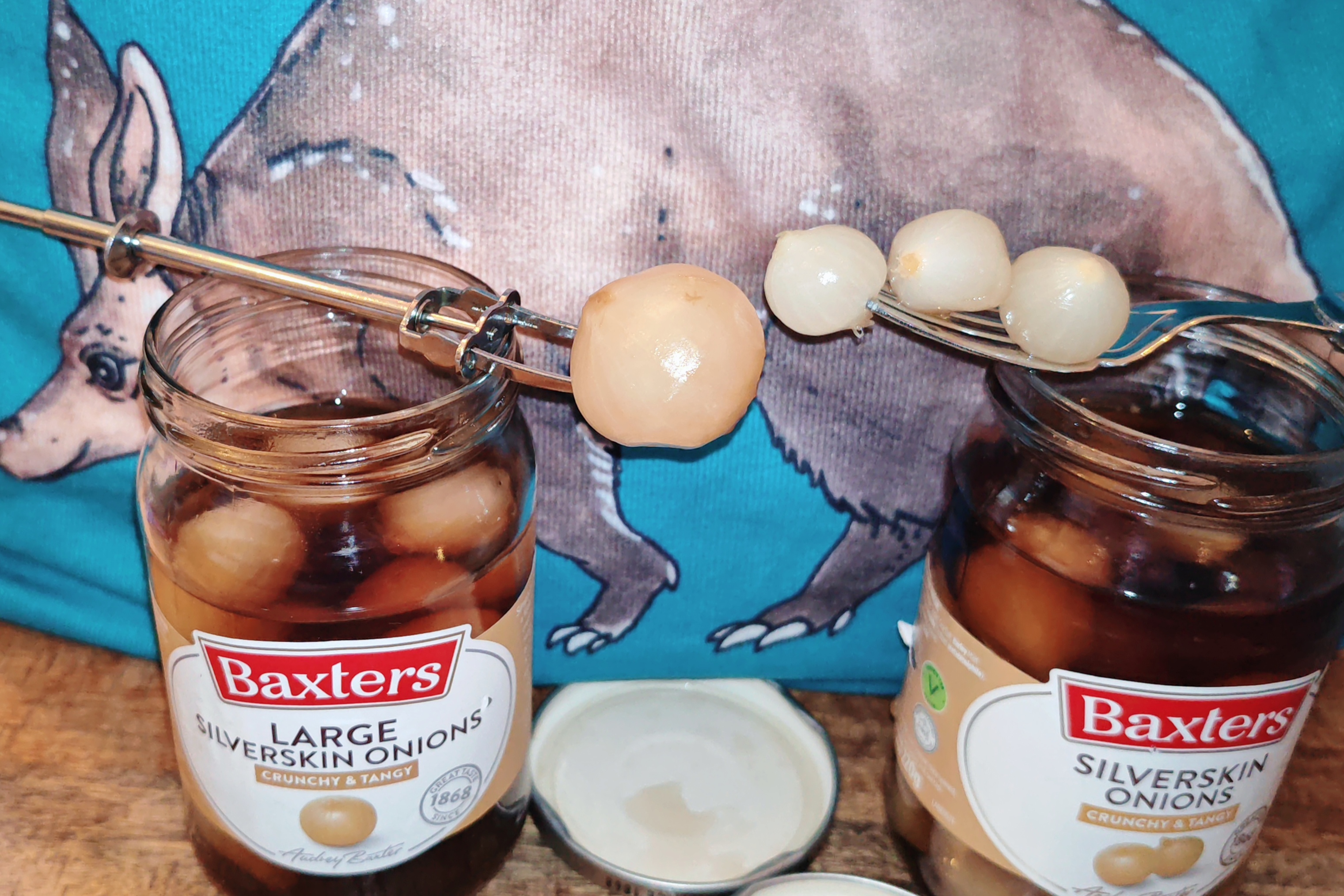 Photo: The testing phase in full swing - metal pickle forks hold several pickled onions above each opened jar for comparison. The onions glisten appetisingly in their amber-coloured brine against the wooden surface, whilst the aardvark print completes this thoroughly British tasting setup.