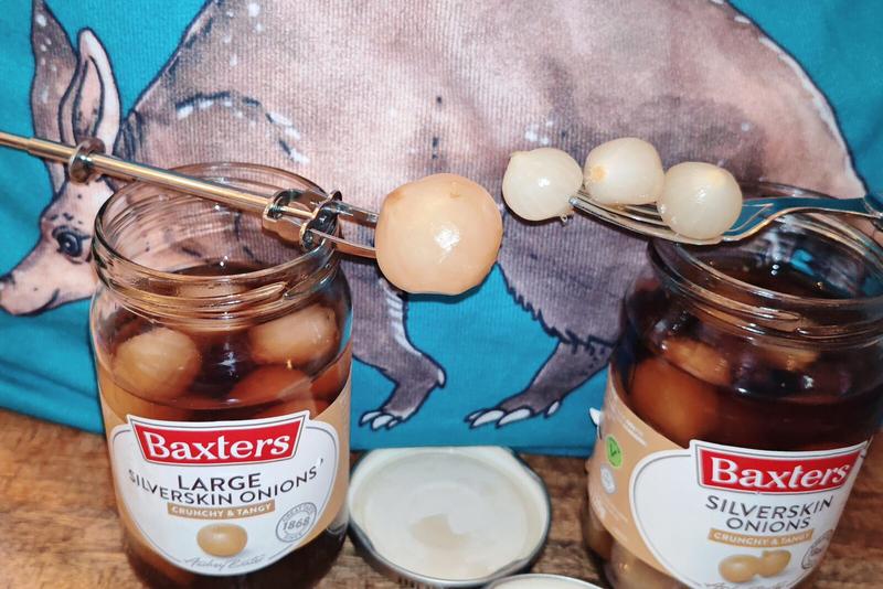 Photo: The testing phase in full swing - metal pickle forks hold several pickled onions above each opened jar for comparison. The onions glisten appetisingly in their amber-coloured brine against the wooden surface, whilst the aardvark print completes this thoroughly British tasting setup.