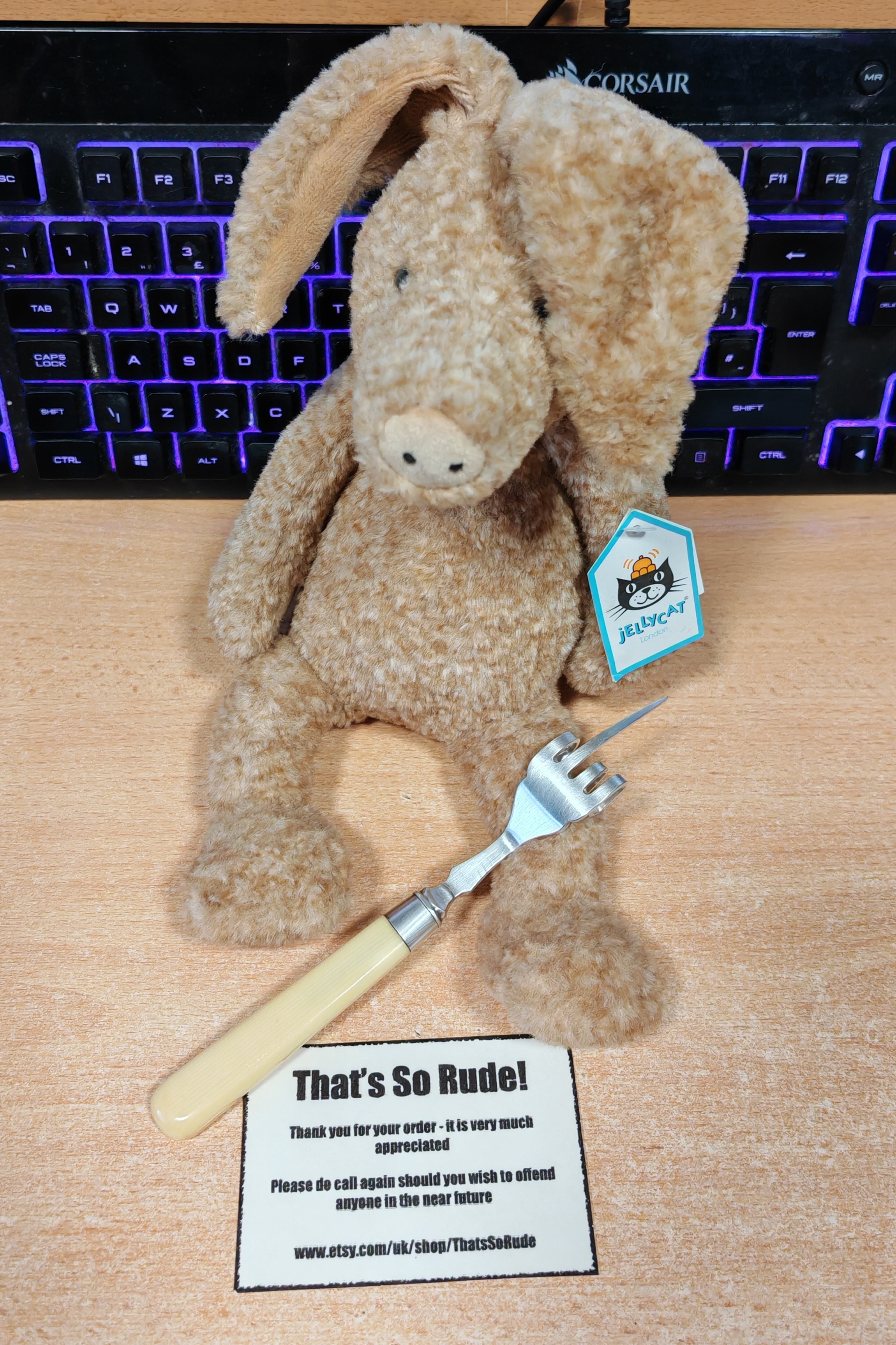 A cream-coloured plush aardvark sits on a wooden desk, brandishing a four-pronged pickle fork where the middle tine has been bent upwards in a rather rude gesture. Behind the aardvark, a keyboard glows with purple backlighting. In front sits a business card reading "That's So Rude! Thank you for your order - it is very much appreciated. Please do call again should you wish to offend anyone in the near future" with the Etsy shop URL. The aardvark sports a turquoise Jellycat tag, clearly demonstrating that even the most refined stuffed animals occasionally need to express themselves in less than polite ways.