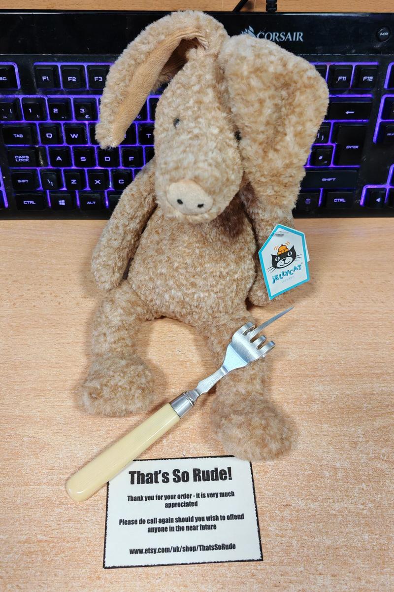 A cream-coloured plush aardvark sits on a wooden desk, brandishing a four-pronged pickle fork where the middle tine has been bent upwards in a rather rude gesture. Behind the aardvark, a keyboard glows with purple backlighting. In front sits a business card reading "That's So Rude! Thank you for your order - it is very much appreciated. Please do call again should you wish to offend anyone in the near future" with the Etsy shop URL. The aardvark sports a turquoise Jellycat tag, clearly demonstrating that even the most refined stuffed animals occasionally need to express themselves in less than polite ways.