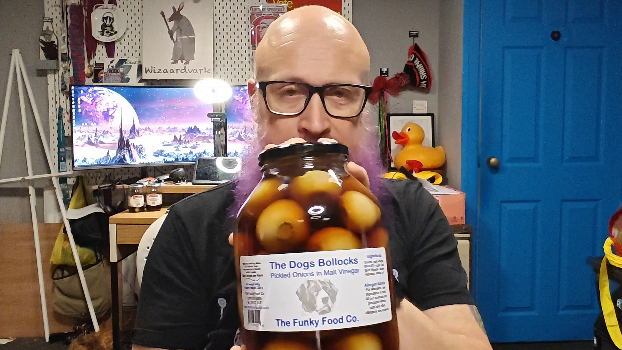 Bald man with glasses holding up a large jar of "The Dogs Bollocks" pickled onions in malt vinegar from The Funky Food Co., featuring a distinguished dog illustration on the label. Behind him is his familiar home office setup with collectibles on shelves, a bright blue door, computer monitors displaying what appears to be a fantasy landscape, and a rubber duck amongst the various pop culture memorabilia. His expression suggests he's about to deliver a verdict on this rather boldly-named preserve.