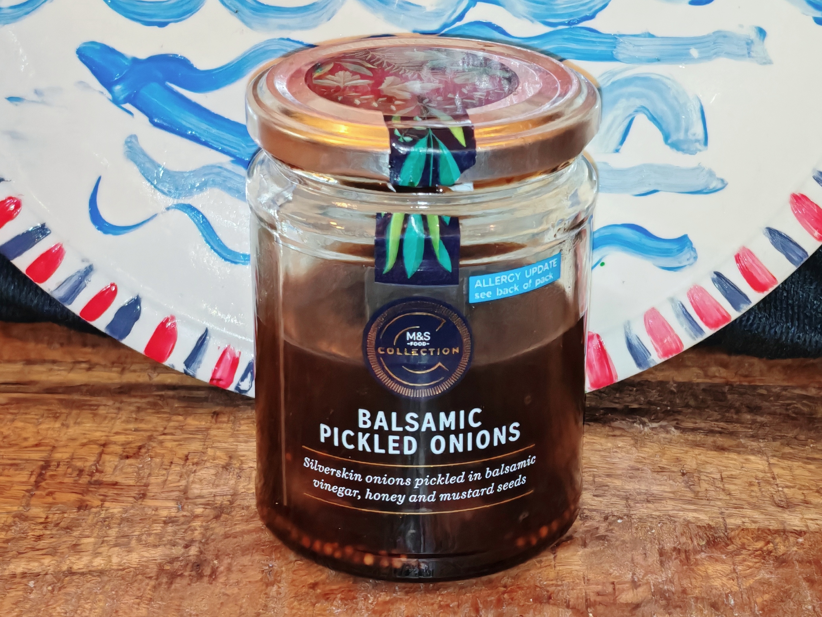 Photo: Full jar of M&S Collection Balsamic Pickled Onions on a wooden surface against a child's hand-painted ceramic plate decorated with blue wavy lines representing the sea and red and white stripes. The dark amber liquid and golden onions are visible through the clear glass, with the premium M&S branding and allergy information clearly displayed on the label.