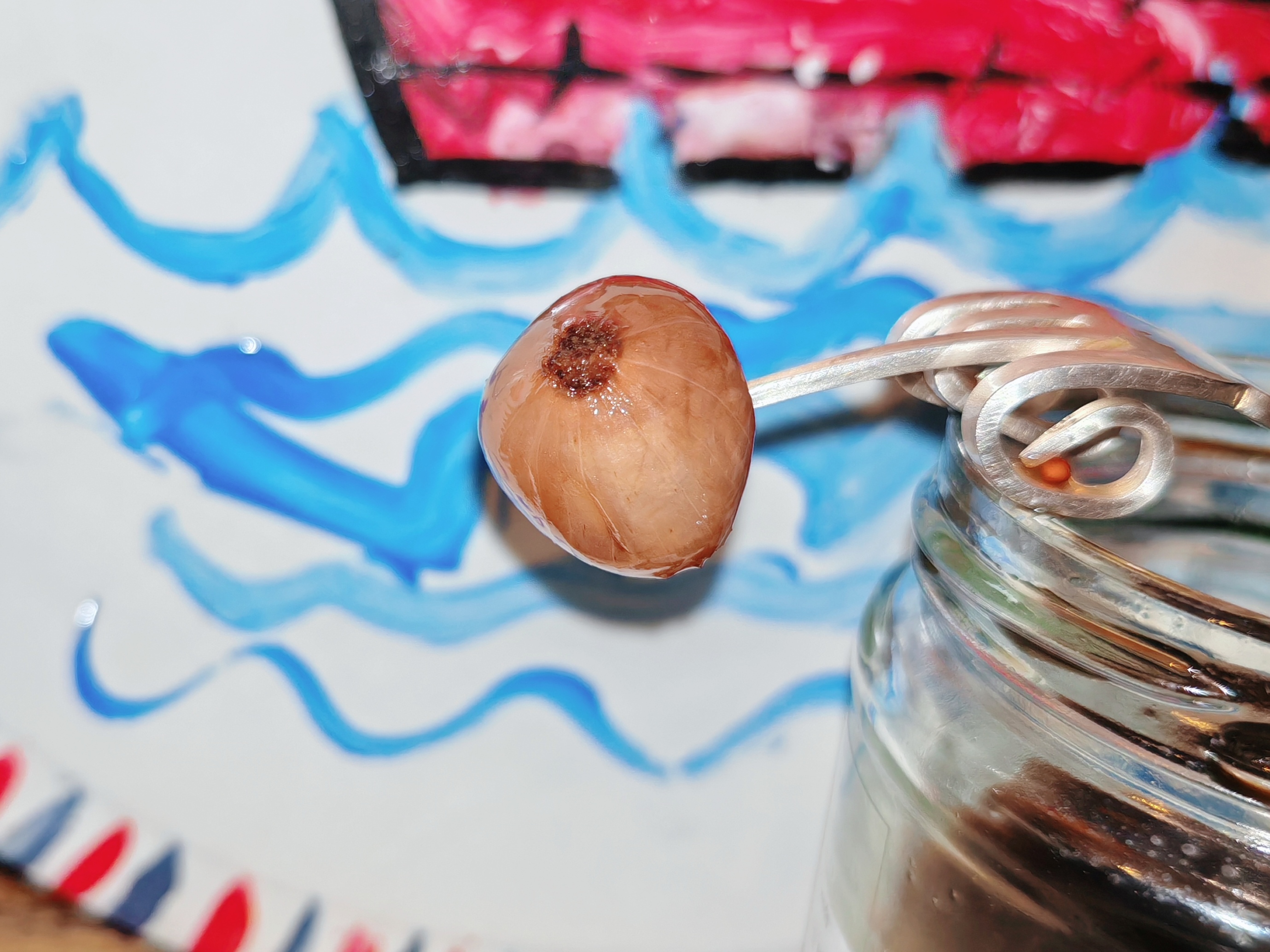 Photo: Single pickled onion on a metal fork against a child's hand-painted ceramic plate decorated with blue wavy lines representing the sea and red and white stripes. The onion appears golden and translucent but darker than most pickled silverskin onions.