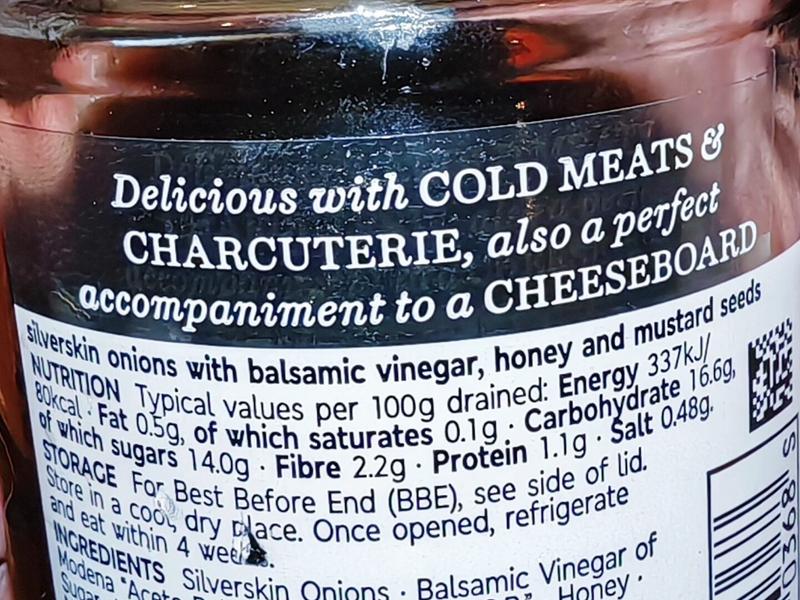 Close-up of a jar label for M&S Collection Balsamic Pickled Onions, showing the product description "Delicious with COLD MEATS & CHARCUTERIE, also a perfect accompaniment to a CHEESEBOARD" along with nutritional information and ingredients list including silverskin onions with balsamic vinegar, honey and mustard seeds.