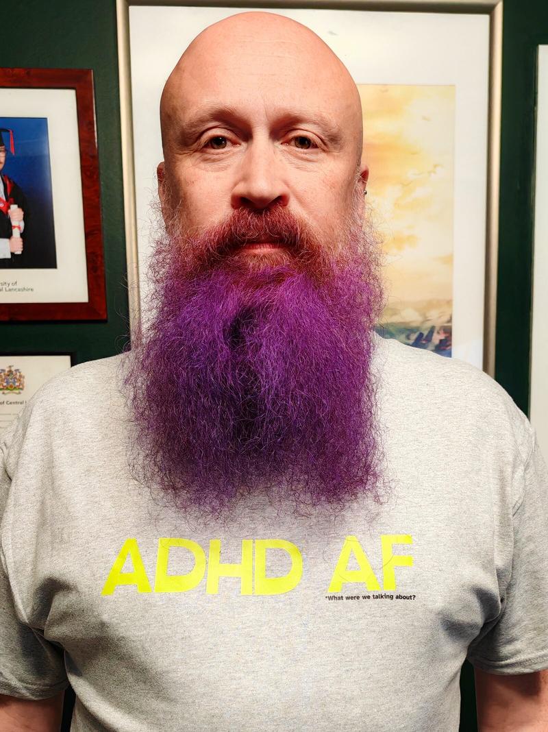 Me sporting my glorious purple beard in front of framed artwork, wearing a grey "ADHD AF" t-shirt with the helpful subtitle "what were we talking about?" Because subtlety is overrated when you're making a point about neurodivergent life.