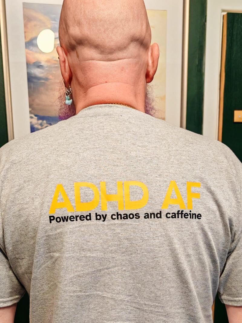 Back view of the same grey t-shirt revealing the punchline: "ADHD AF - Powered by chaos and caffeine." Also visible: the back of my magnificently bald head, a small earring, and the edge of said purple beard.