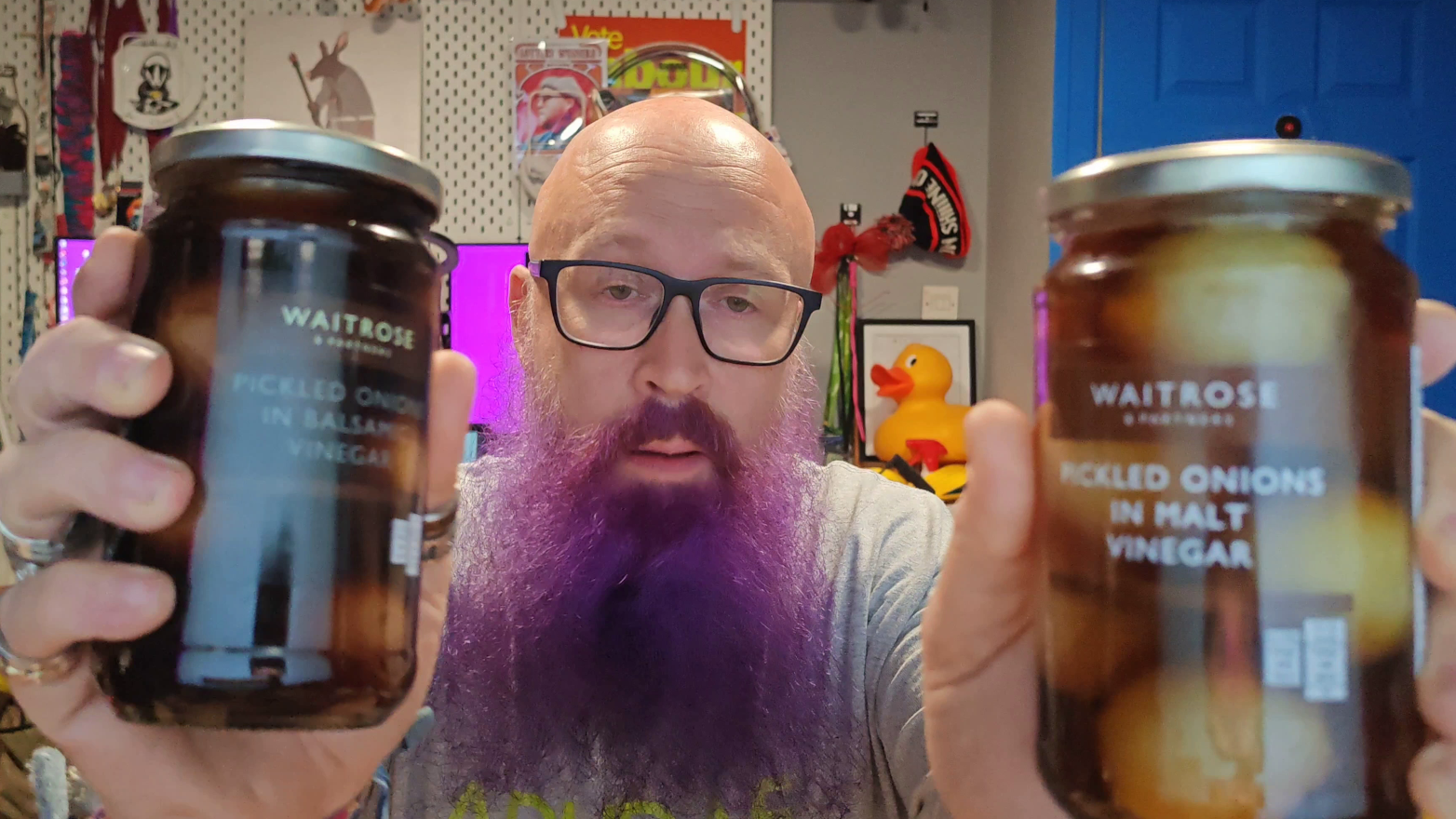 Photo: Bald man with glasses and a purple beard wearing a grey t-shirt, holding up two jars of Waitrose pickled onions - one appears to be pickled onions in balsamic vinegar (left jar with darker contents) and one pickled onions in malt vinegar (right jar with lighter, more traditional-looking onions). Behind him is his familiar home office setup with shelves of collectibles, a bright blue door, and a rubber duck amongst various pop culture memorabilia. His expression suggests he's mid-comparison between these two Waitrose offerings.