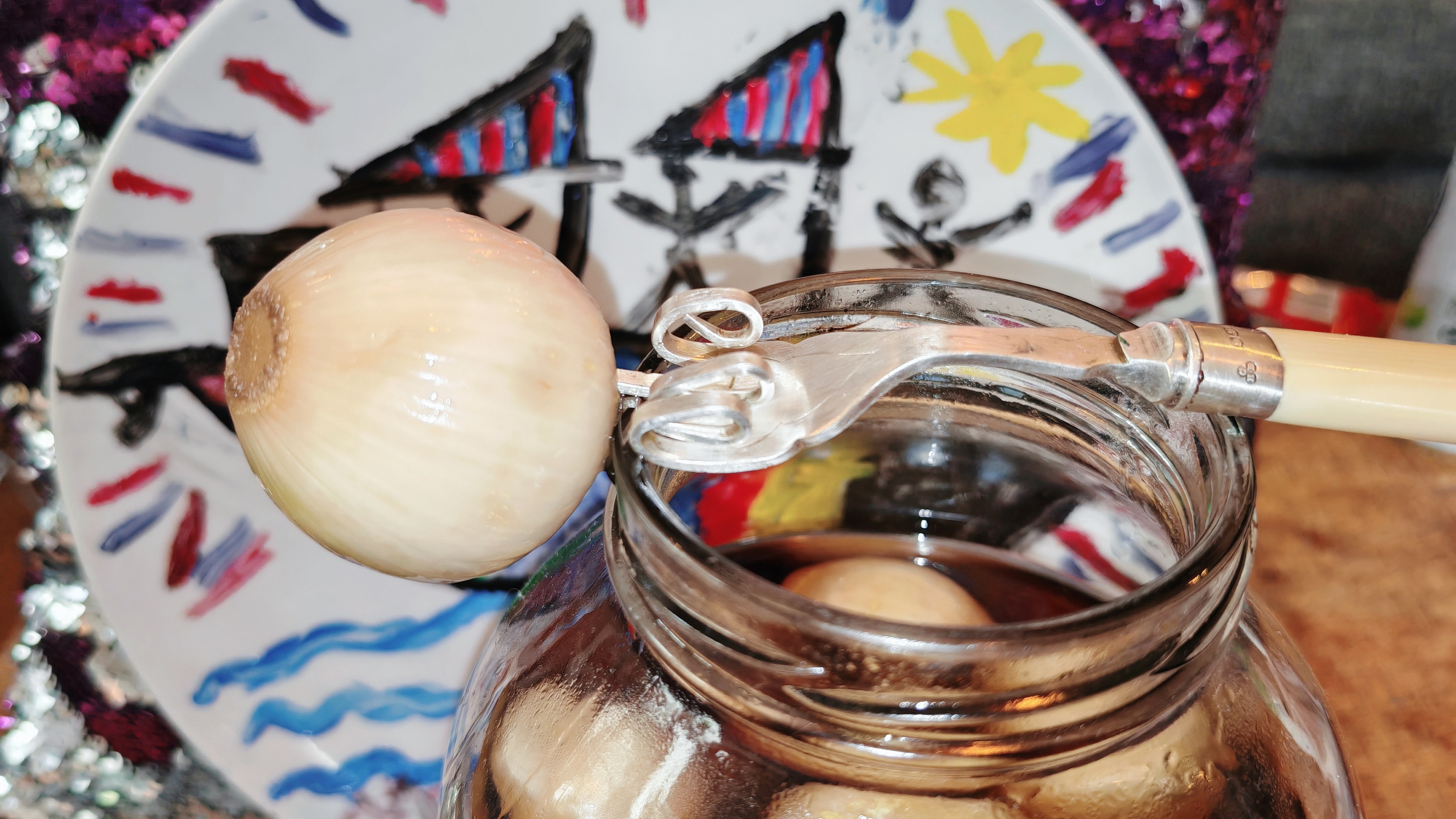 Photo: Close-up of an open jar of pickled onions with a single pale onion balanced on a cocktail stick across the rim. Behind the jar sits the same decorative plate with its cheerful red, blue and yellow design, whilst magenta sequins sparkle in the background.
