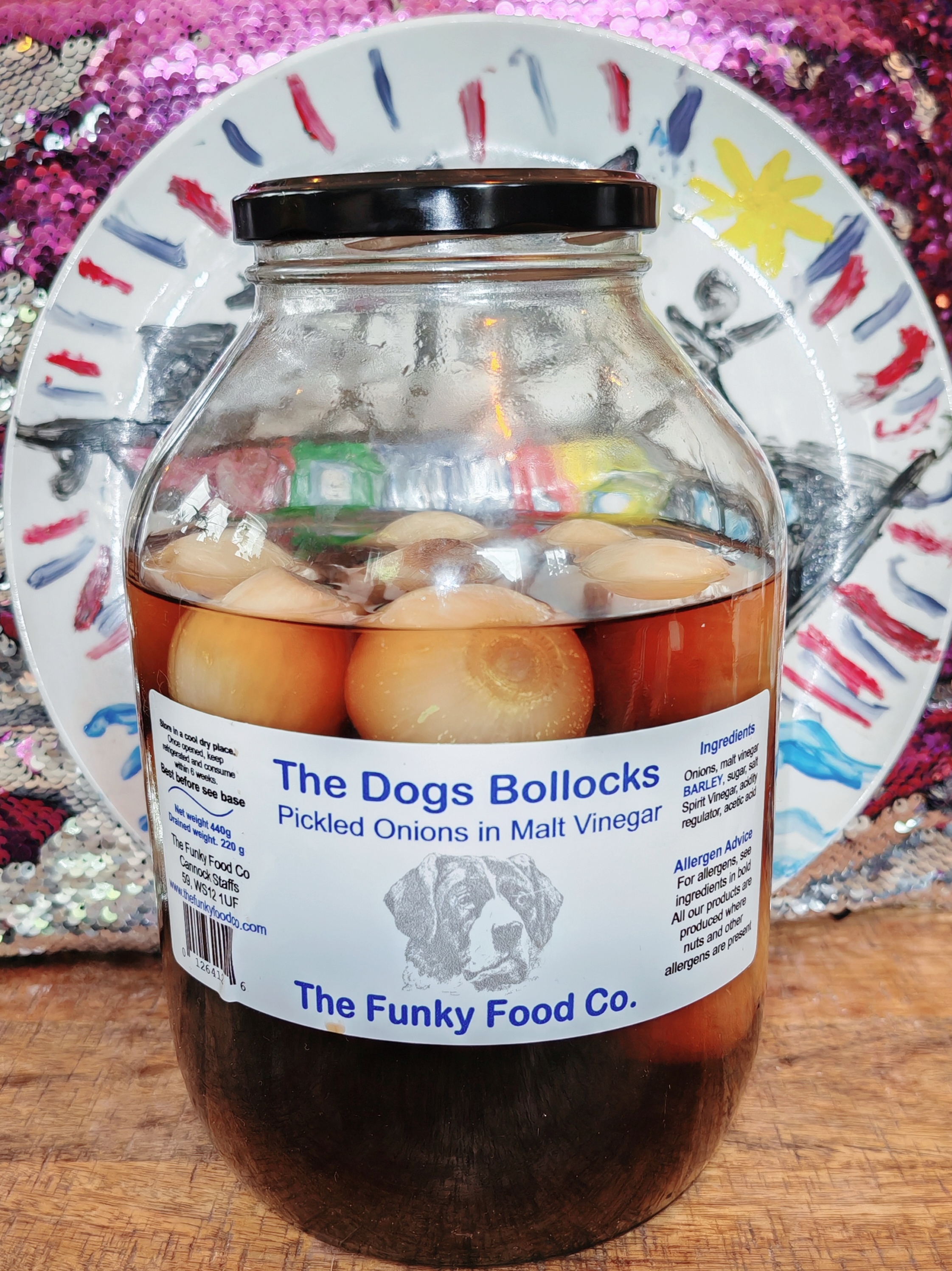 Photo: A large glass jar of pickled onions in malt vinegar sits on a wooden surface. The label reads "The Dogs Bollocks Pickled Onions in Malt Vinegar" from The Funky Food Co., featuring a sketch of a dog's face. Behind the jar is a white plate decorated with red and blue brushstrokes and a yellow flower. The background shows a shimmery magenta and silver sequined fabric.