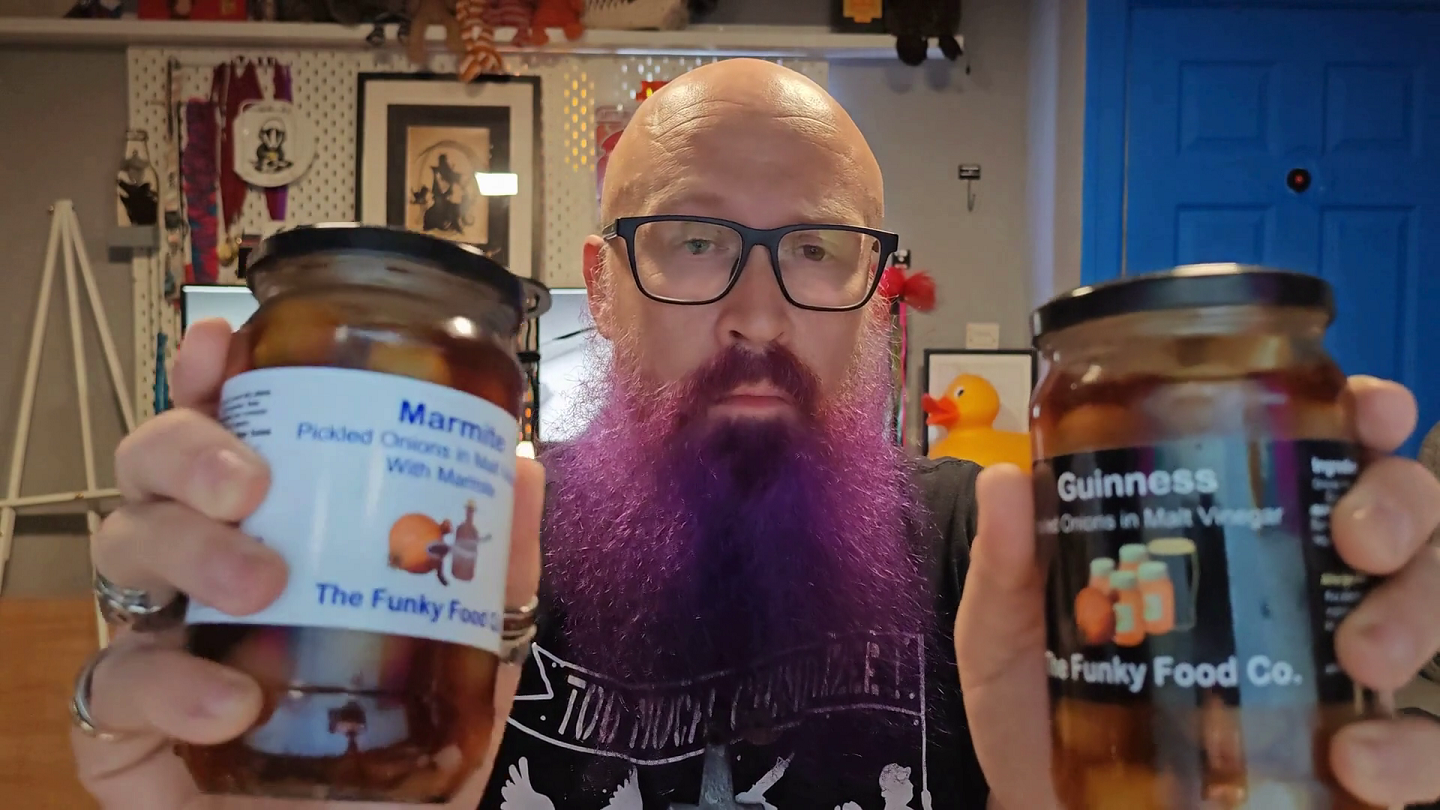Bald man with glasses and a purple beard wearing a black t-shirt, holding up two jars of Funky Food Co. pickled onions - Marmite pickled onions in malt vinegar (left jar with white label featuring the distinctive Marmite branding) and Guinness pickled onions in malt vinegar (right jar with black label). Behind him is his familiar home office with shelves of collectibles, framed artwork, a bright blue door, and a rubber duck visible amongst the memorabilia. His expression suggests he's about to embark on what could be described as either a culinary adventure or an act of madness, depending on one's tolerance for bold flavours.