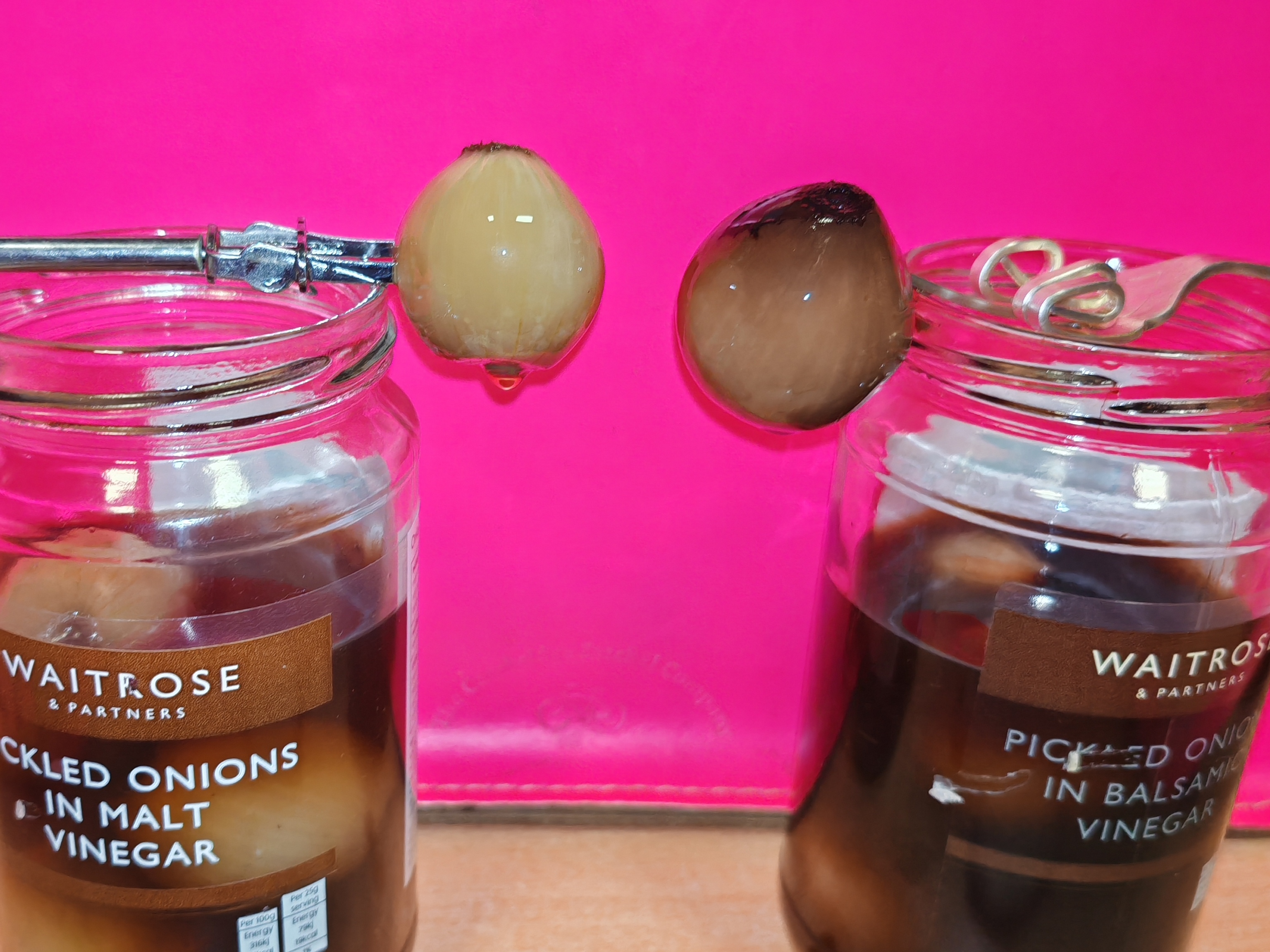 Photo: Two pickled onions suspended on pickle forks between the opened jars against the pink background. The left onion from the malt vinegar is pale cream-coloured, the right one from balsamic vinegar is darker brown. A compelling study in allium contrast.