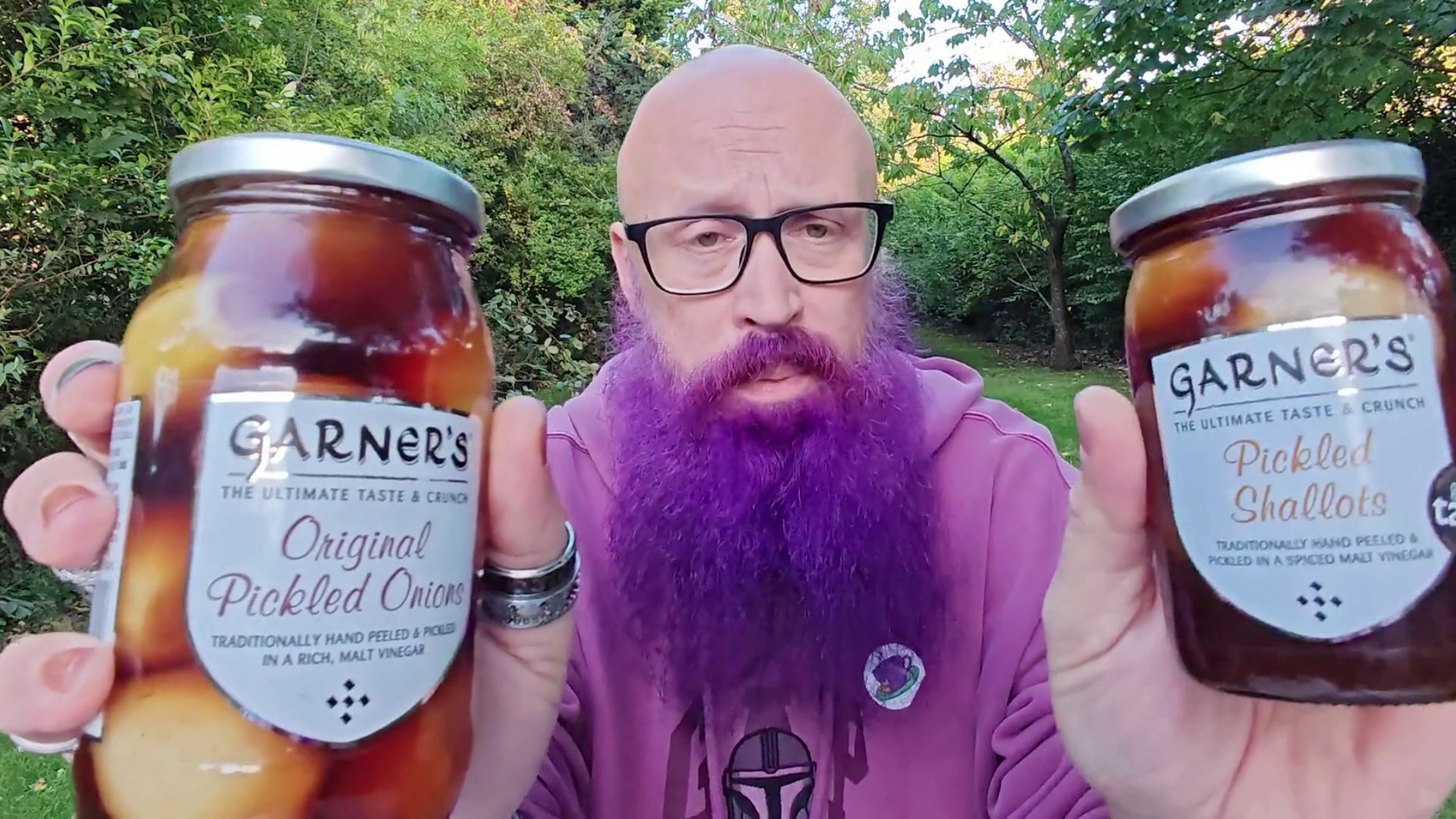 Bald man with glasses and a vibrant purple beard wearing a purple hoodie, outdoors in a garden and holding up two jars of Garner's pickled onions - "Original Pickled Onions" (traditionally hand peeled and pickled in a rich malt vinegar) on the left, and "Pickled Shallots" (traditionally hand peeled and pickled in a spiced malt vinegar) on the right. Behind him is lush green foliage with trees and shrubs creating a natural backdrop. His expression suggests he's about to deliver his considered opinion on these artisanal preserves.