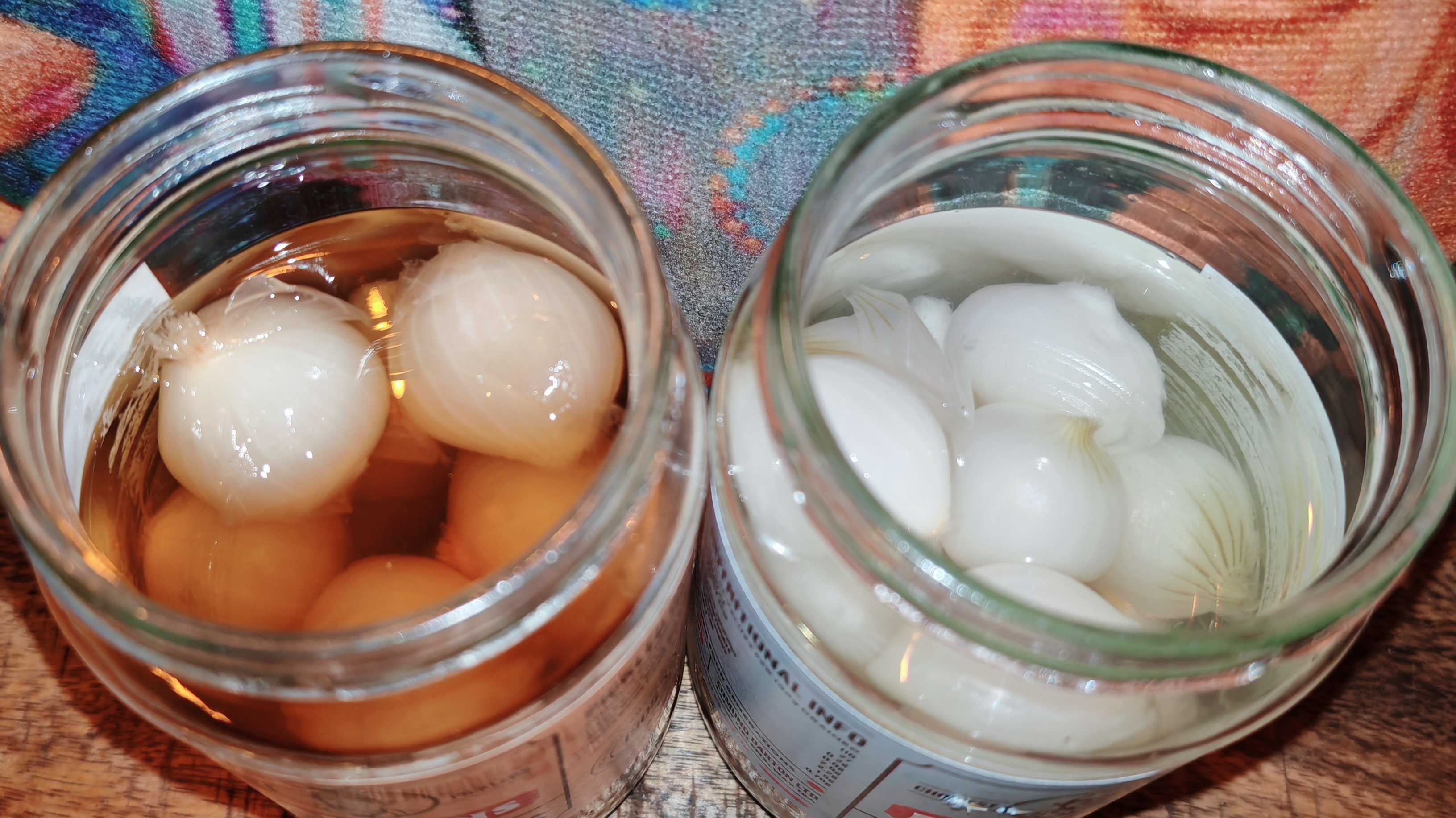 Overhead view of two opened jars on a colourful patterned fabric. Left jar contains traditional pickled onions in brown malt vinegar. Right jar contains silverskin onions in clear vinegar. Both show whole, peeled onions submerged in their respective pickling liquids.