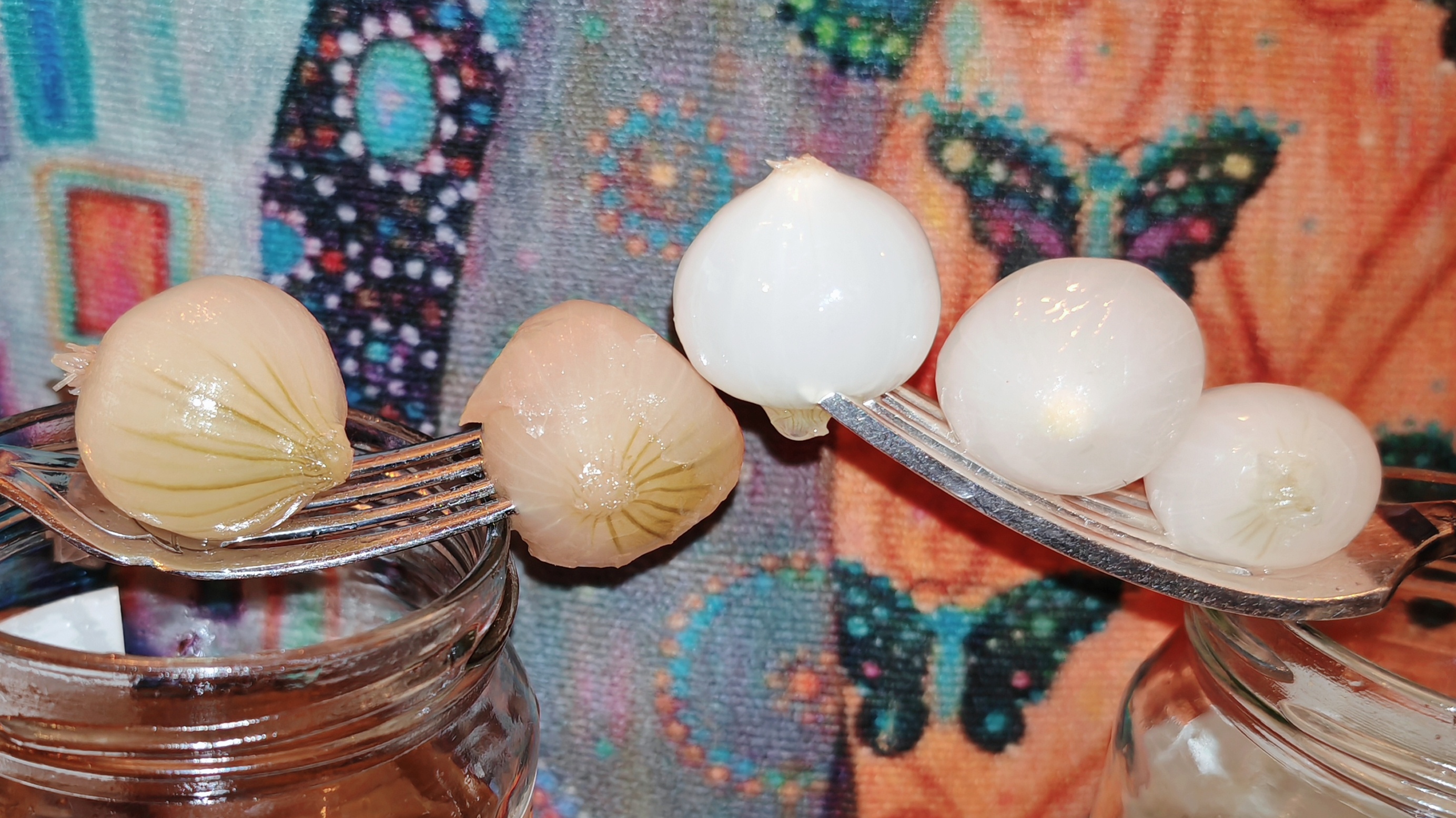  Two forks extended over the opened jars, each holding pickled onions. Left fork holds two pale brown traditional pickled onions with visible root marks. Right fork holds three white silverskin onions. Colourful patterned fabric in background.