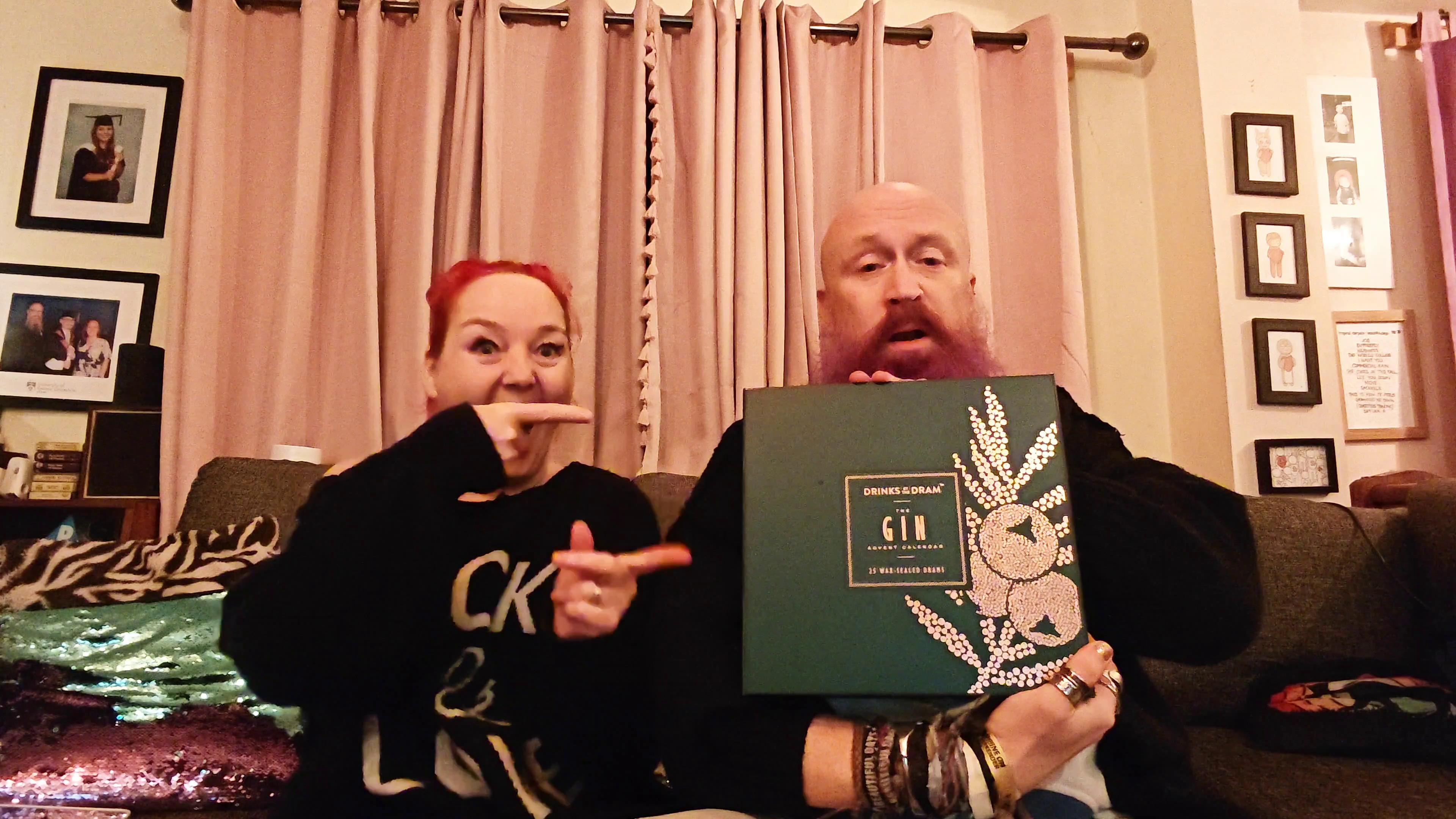 Photo: Two people sitting on a sofa in a living room. On the left, a woman with bright red hair wearing a black top, smiling at the camera. On the right, a bald man with a purple beard wearing a black t-shirt with "GAP" printed on it, looking directly at the camera with a neutral expression. Behind them are pale pink curtains and a wall with framed photographs. In the foreground on the left is part of a decorative cushion with an animal print pattern.