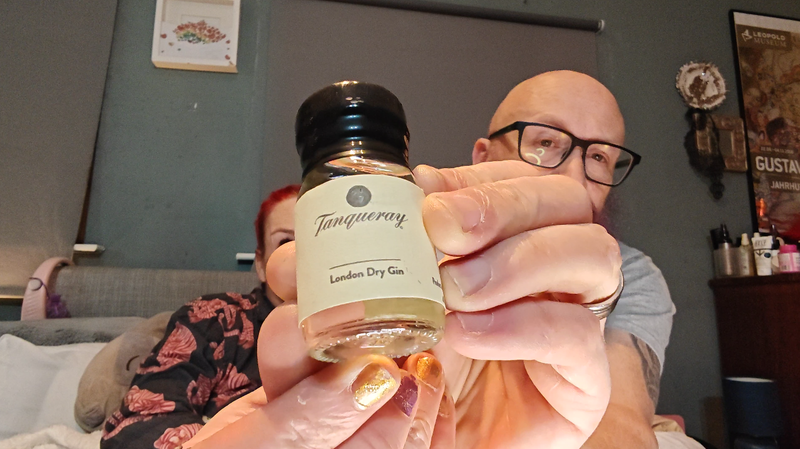 Photo: A bald man with black-framed glasses and a full purple beard holds a small glass bottle of Tanqueray London Dry Gin towards the camera. The bottle has a dark green cap and a cream-coloured label with gold lettering. His hands dominate the foreground, with gold-painted nails visible on one hand. Behind him, a woman with bright red hair wearing a black top with coral or pink floral patterns sits on a bed with a grey fabric headboard. The bedroom setting features a grey wall with a white-framed artwork showing colourful butterflies or flowers in the upper left, and a Leopold Museum poster on the right side.
