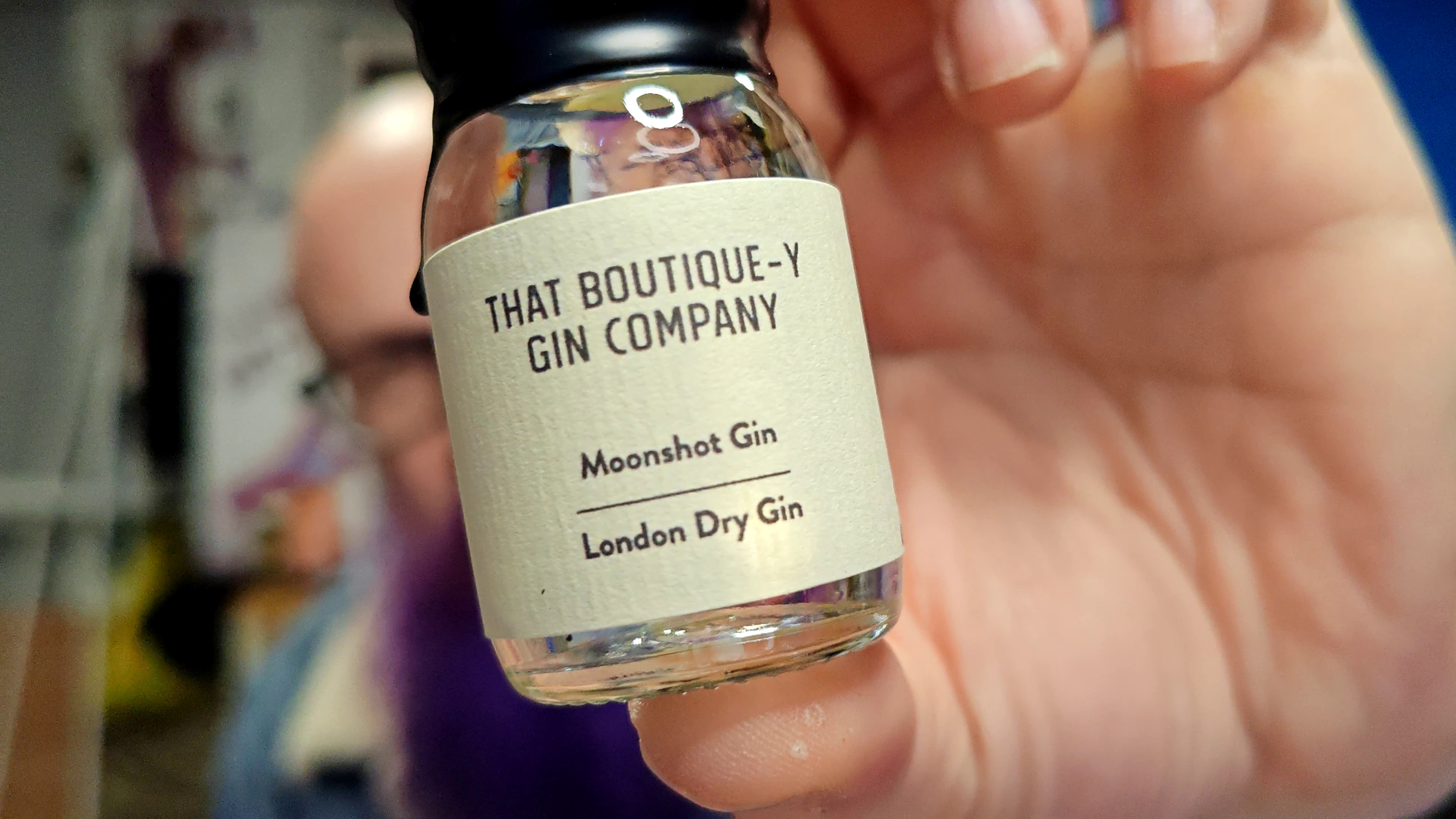 Photo: A hand holding a small glass bottle of gin in sharp focus towards the camera, with a blurred background showing a person. The bottle has a black screw cap and a cream-coloured label reading "THAT BOUTIQUE-Y GIN COMPANY" at the top, followed by "Moonshot Gin" and "London Dry Gin" below. The clear liquid is visible inside the bottle. The person holding it is out of focus but appears to be wearing something pink or peach-coloured. In the soft-focus background, hints of a room with various colours including purple and yellow are visible.