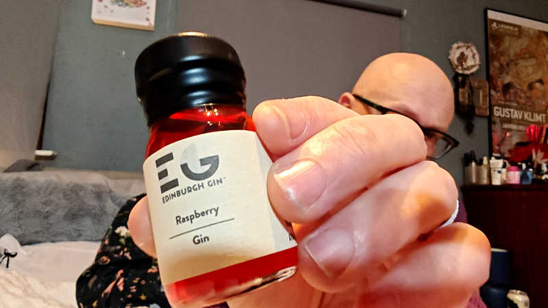 Photo: A hand holding a small glass bottle of Edinburgh Gin towards the camera in sharp focus. The bottle contains red liquid and has a black wax sealed cap. The cream-coloured label displays the Edinburgh Gin logo at the top, followed by "Raspberry" and "Gin" in black text. Behind the bottle, slightly out of focus, a bald man wearing black-framed glasses sits in a bedroom setting. The grey wall features a white-framed artwork showing colourful butterflies in the upper left, and a Leopold Museum poster showing Gustav Klimt artwork on the right. A grey fabric headboard and white bedding are visible, along with a woman in a black floral top partially visible to the left.