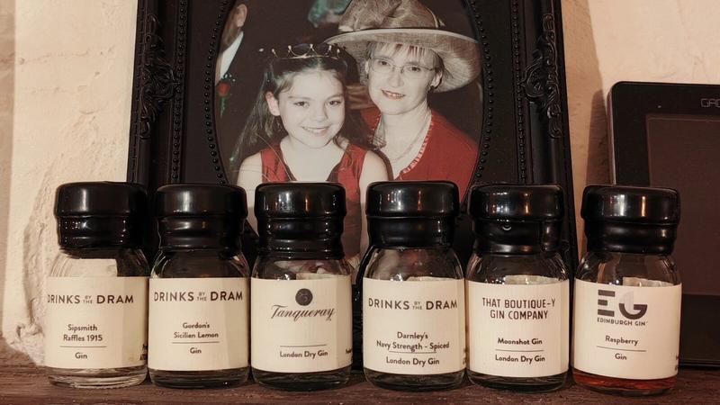 Photo: Six empty small glass bottles of gin arranged in a row on a dark wooden surface, displayed in front of a framed photograph. Each bottle has a black screw cap and cream-coloured label. From left to right, the labels read: "DRINKS BY THE DRAM Sipsmith Raffles 1915 Gin", "DRINKS BY THE DRAM Gordon's Sicilian Lemon Gin", "Tanqueray London Dry Gin", "DRINKS BY THE DRAM Darnley's Navy Strength - Spiced London Dry Gin", "THAT BOUTIQUE-Y GIN COMPANY Moonshot Gin London Dry Gin", and "EDINBURGH GIN Raspberry Gin". Behind the bottles sits an ornate dark wooden frame with decorative carved corners containing a 25-year-old photograph of an 8-year-old girl wearing a small crown and red dress standing next to a woman wearing a wide-brimmed hat and red top. The scene is set against a textured cream or beige wall, with part of a dark electronic device visible on the right edge.