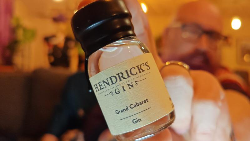 A hand holding a small glass bottle of gin towards the camera in sharp focus against a warm, softly lit background. The bottle has a black wax sealed screw cap and a cream-coloured label reading "HENDRICK'S GIN" in large text at the top, with "DISTILLED AND BOTTLED IN SCOTLAND" in smaller text above, followed by "Grand Cabaret" and "Gin" below. The bottle contains pale golden liquid. The background is deliberately blurred, showing warm amber and yellow lighting from string bulbs, with indistinct figures and pink fabric visible. The overall lighting creates a cosy, intimate atmosphere.