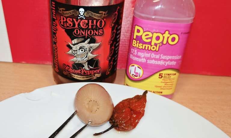 Photo: White plate with two items - a pale pickled onion on a fork on the left showing visible onion rings, and a wrinkled dark red ghost pepper on the right. Behind them, the Psycho Onions jar and a pink bottle of Pepto Bismol stomach relief medicine. The compositional joke is clear.
