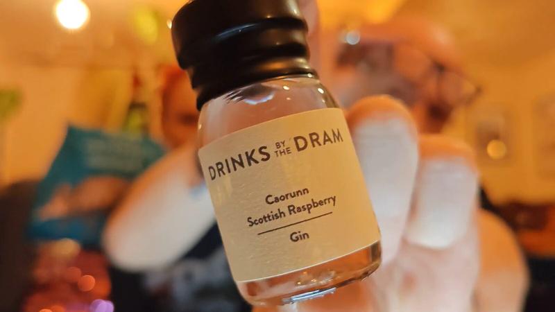 focus against a warm, softly blurred background. The bottle has a black screw cap and a cream-coloured label reading "DRINKS BY THE DRAM" at the top, followed by "Caorunn Scottish Raspberry" and "Gin" below. The bottle contains pale pink or rose-tinted liquid. The background is deliberately out of focus, showing warm yellow and orange lighting, with a teal-coloured cushion visible on the left and indistinct figures in the centre-right. Warm string lights create bokeh effects in the background.