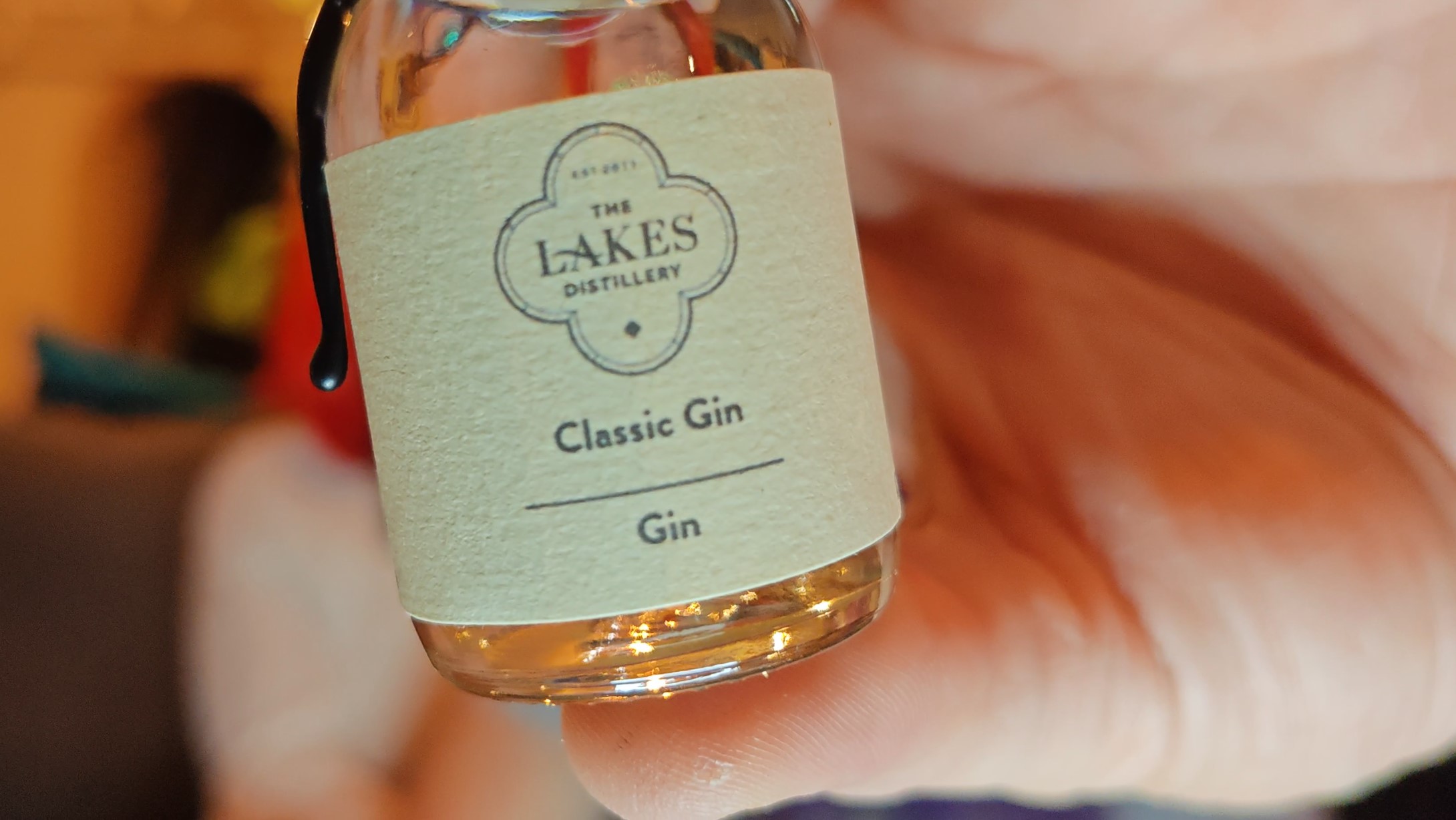 Photo: A hand holding a small glass bottle of gin in sharp focus against a soft, blurred background with warm peachy-pink tones. The bottle has a black screw cap and a pale mint green or cream-coloured label with "THE LAKES DISTILLERY" logo at the top in a cloud-like decorative border, followed by "Classic Gin" and "Gin" below. The bottle contains pale golden liquid. The background is deliberately out of focus, showing warm lighting and indistinct shapes suggesting people and interior furnishings.